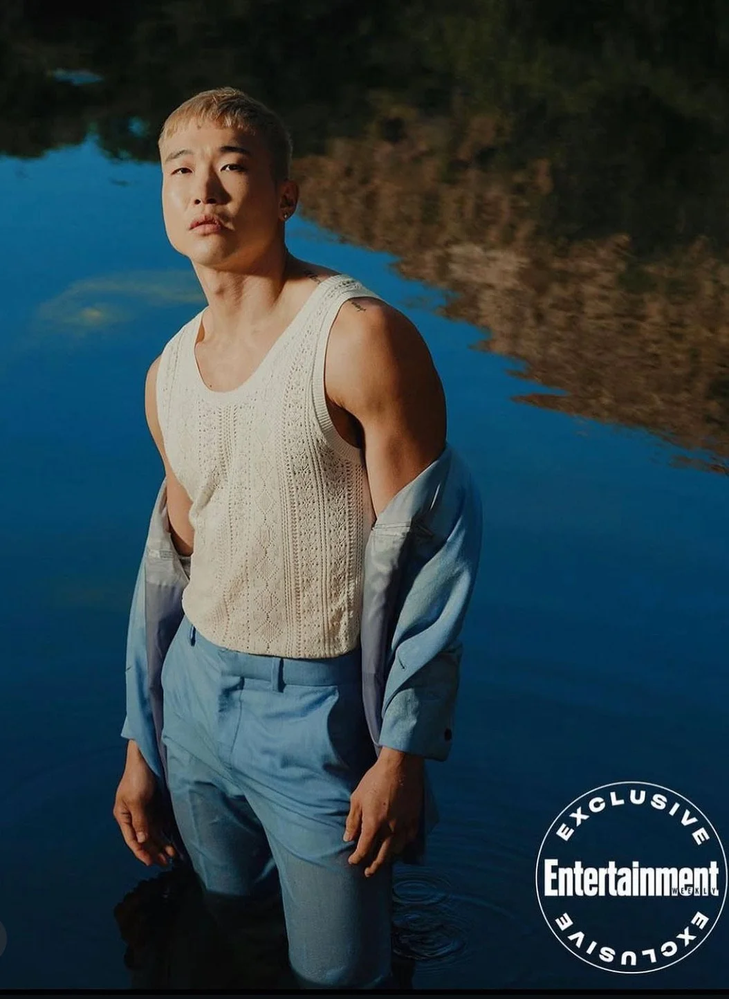A person standing in a body of water at sunset, wearing a sleeveless cream-colored top, light blue pants, and a partially unzipped light blue jacket, with a reflection of the landscape and sky in the water.