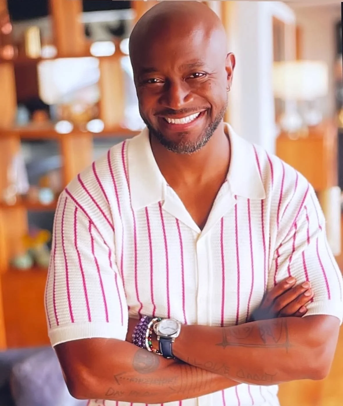 A smiling man with a shaved head and beard, wearing a white and pink striped polo shirt, with his arms crossed, standing indoors with wooden shelves and blurred decor in the background.