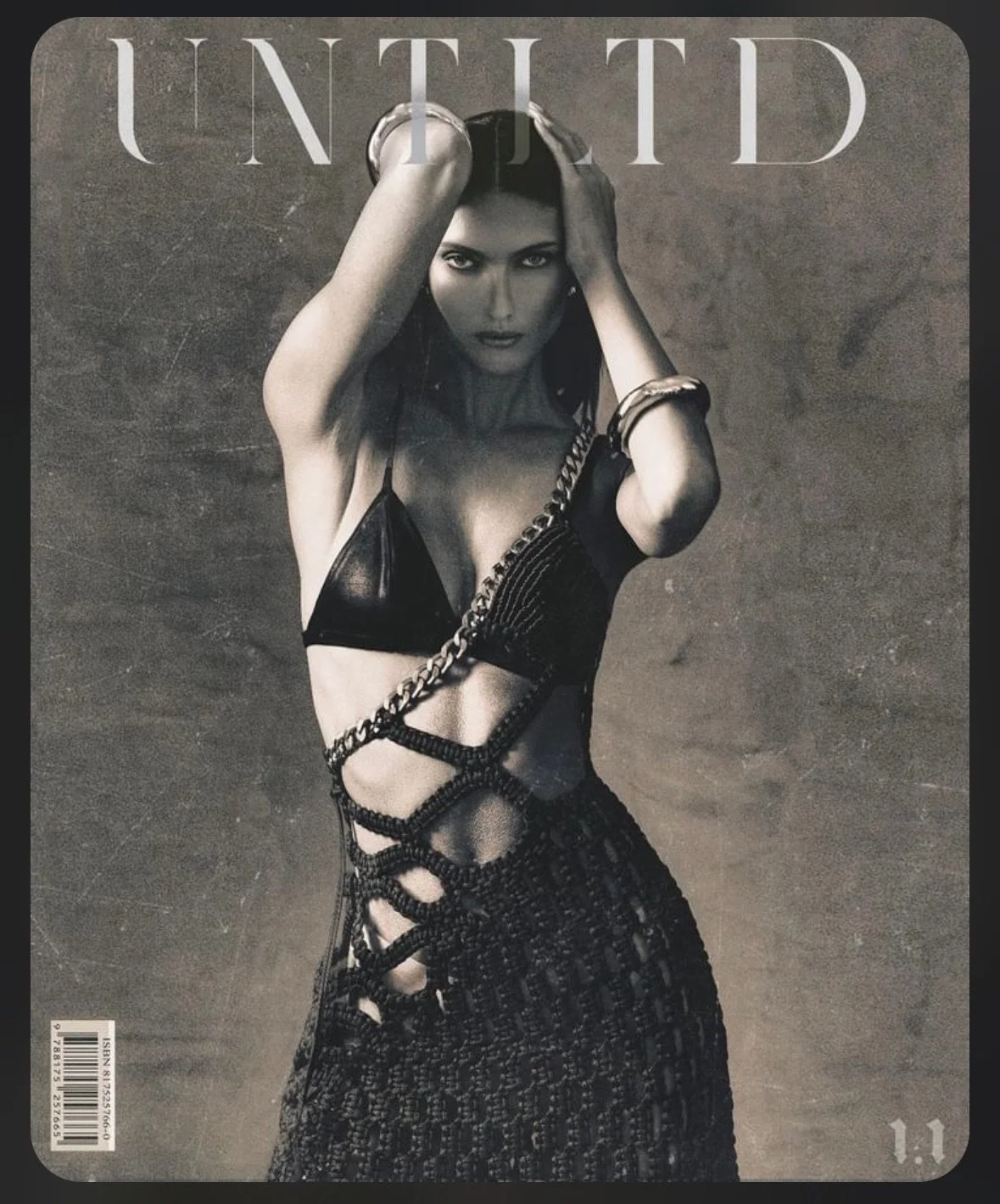 Black and white photo of a woman posing against a plain background with her hands on her head. She is wearing a revealing outfit with a chain accessory, including a bikini-style top and a skirt with intricate open lattice design. The word 'UNITED' is