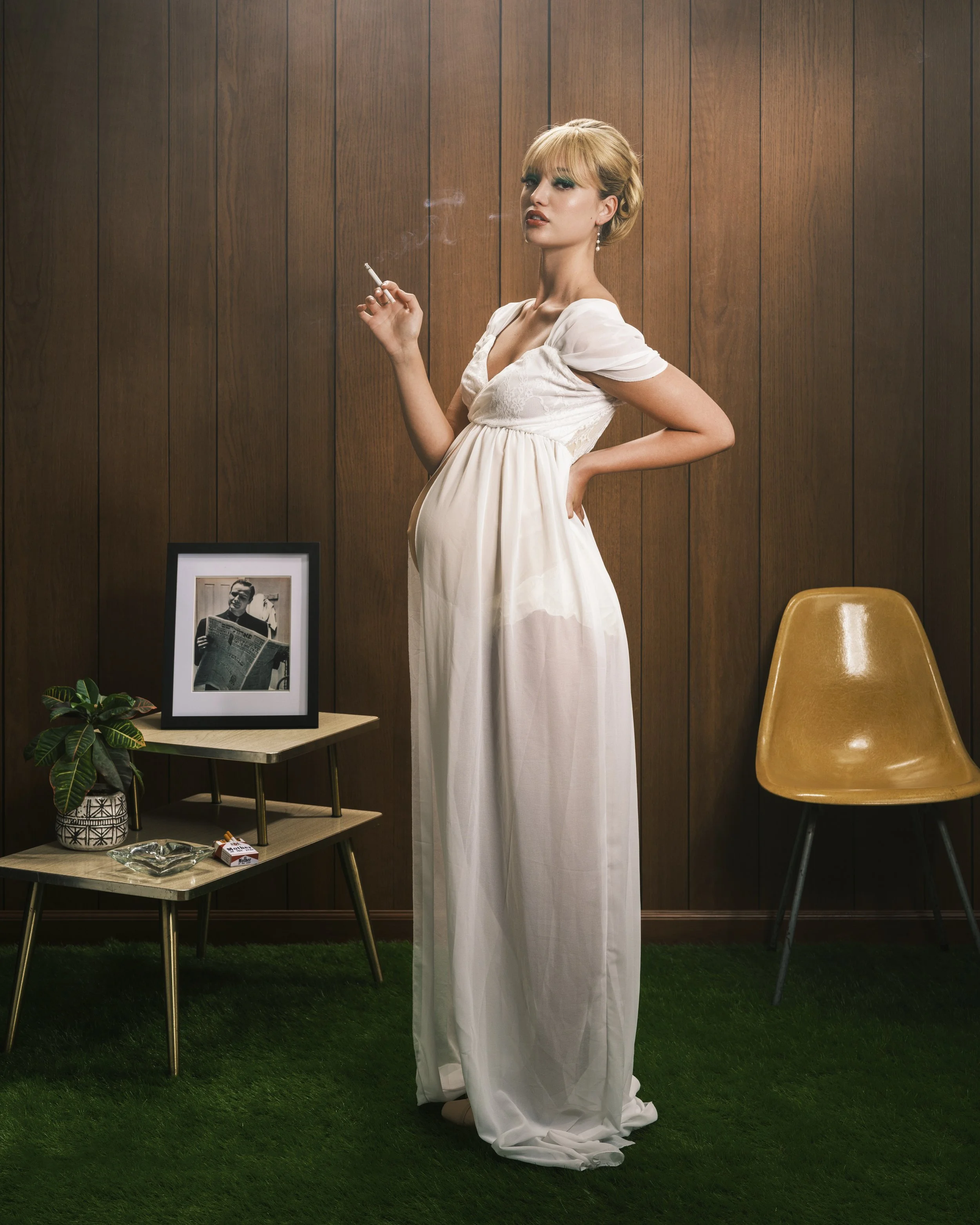 A woman in a white dress stands in a room with wood-paneled walls, smoking a cigarette. There is a framed black-and-white photograph on a small table, along with a potted plant, a pack of cigarettes, and an ashtray. A mustard-colored chair is next to