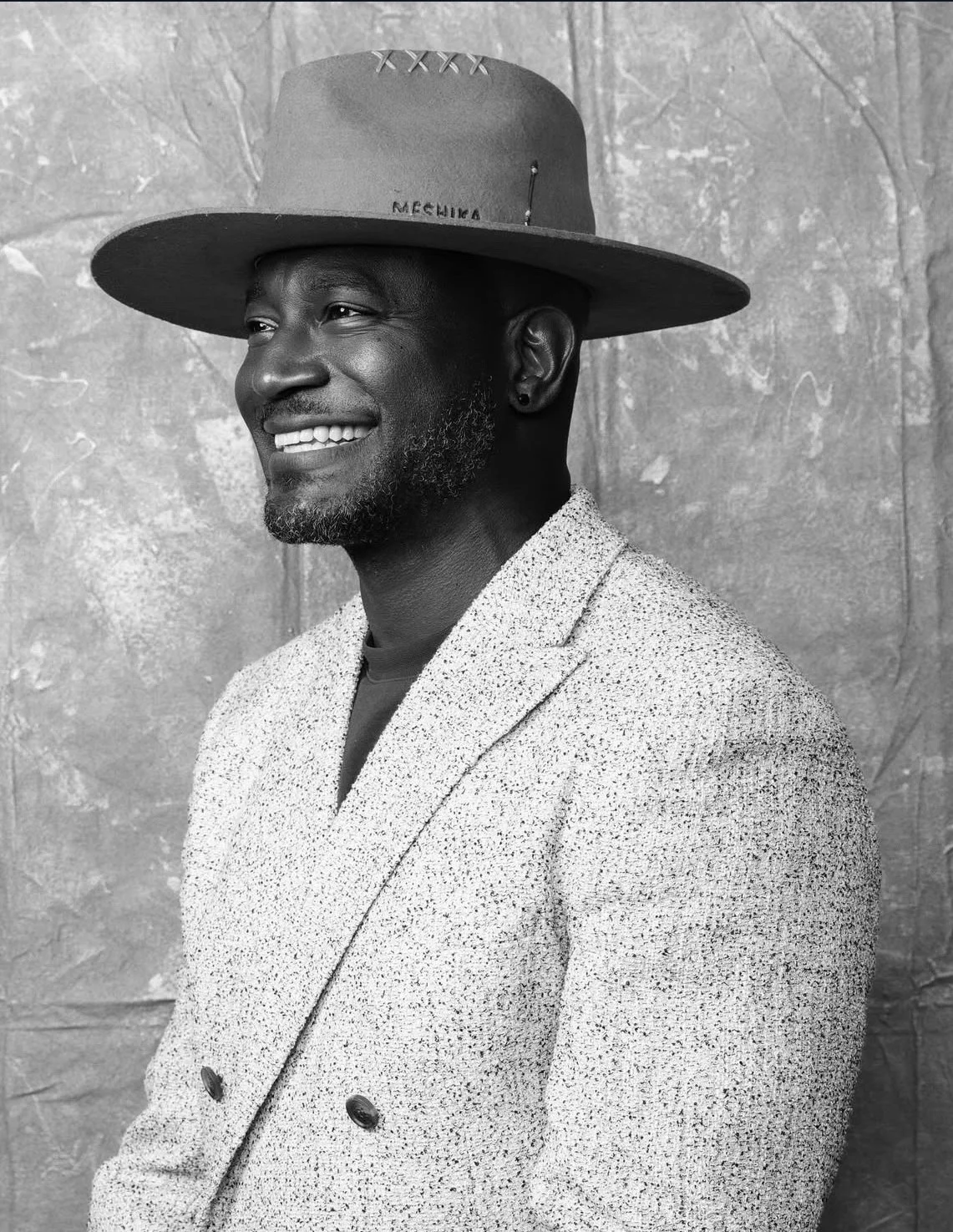A black and white portrait of a man wearing a textured blazer and a wide-brimmed hat, smiling and looking to the side, standing against a plain textured background.