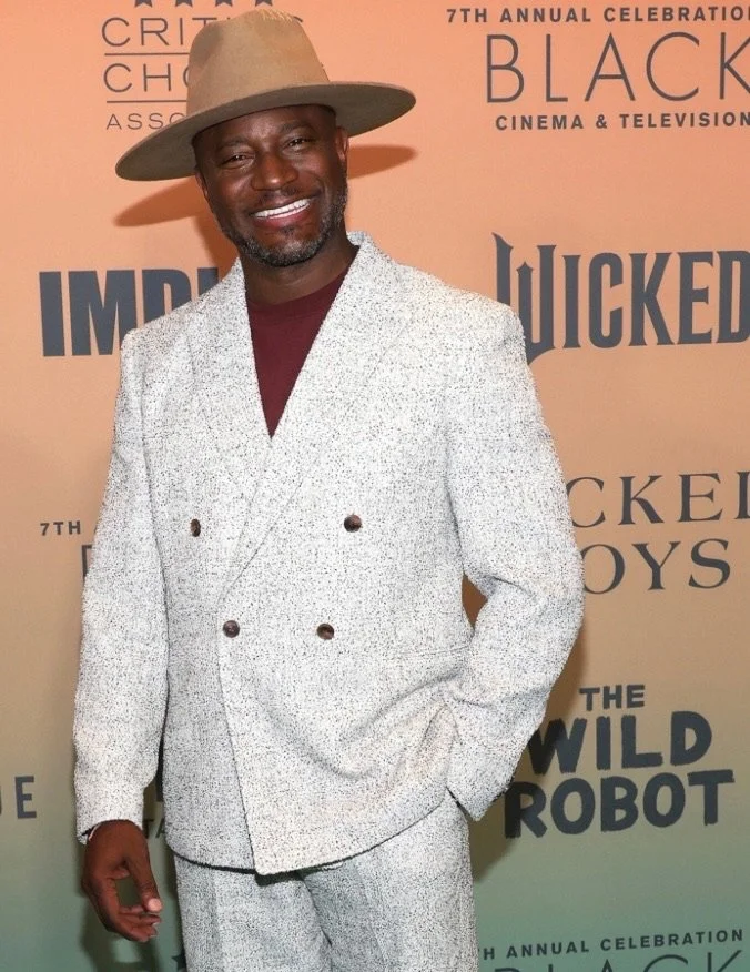 A man in a light-colored, textured double-breasted suit and a matching wide-brimmed hat, smiling at a 7th annual celebration event with a pinkish-beige backdrop featuring black text and logos.