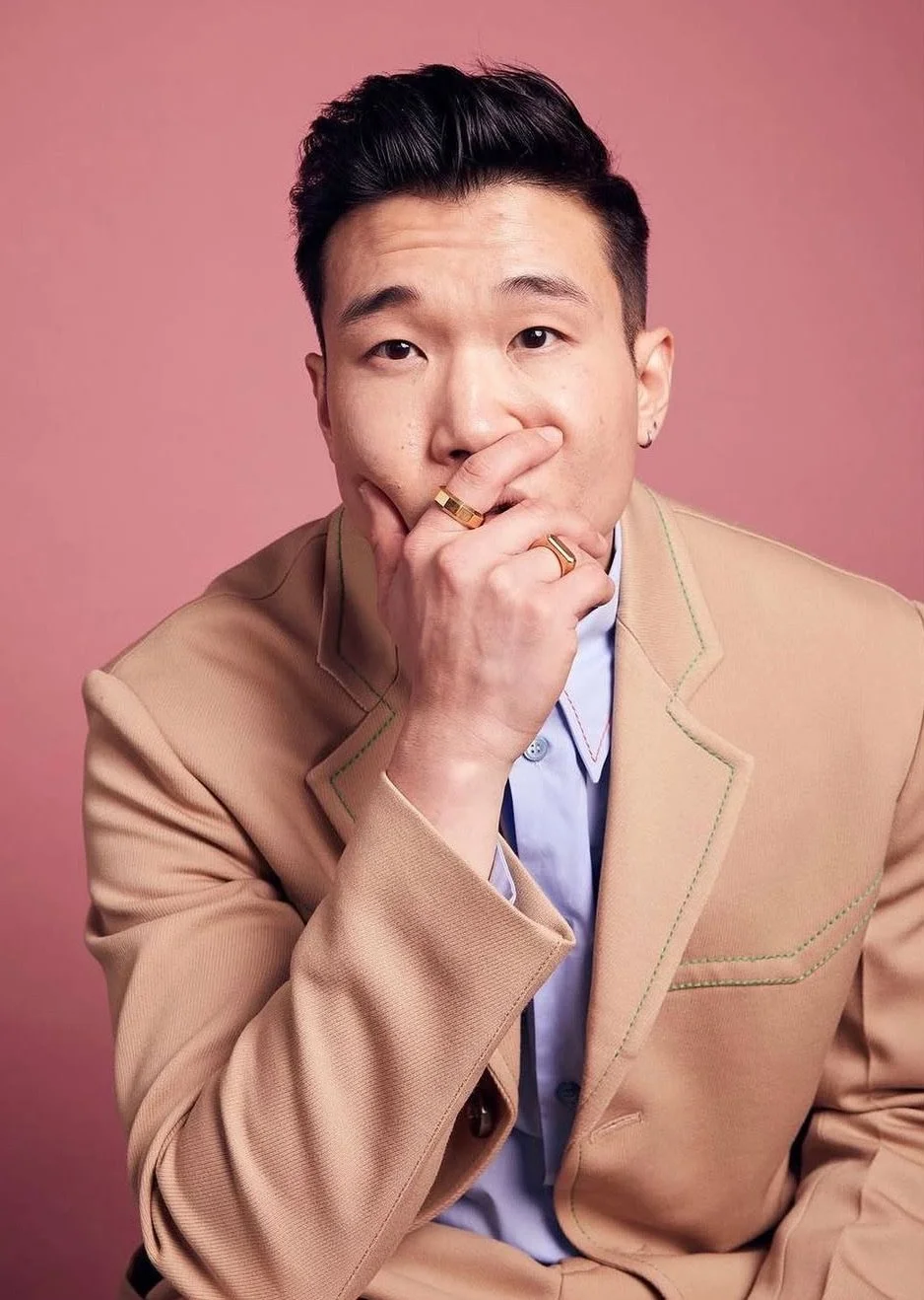 A young man with dark hair, wearing a beige blazer and light blue shirt, posed against a pink background. He is resting his chin on his hand, with rings on his fingers, and looks directly at the camera.