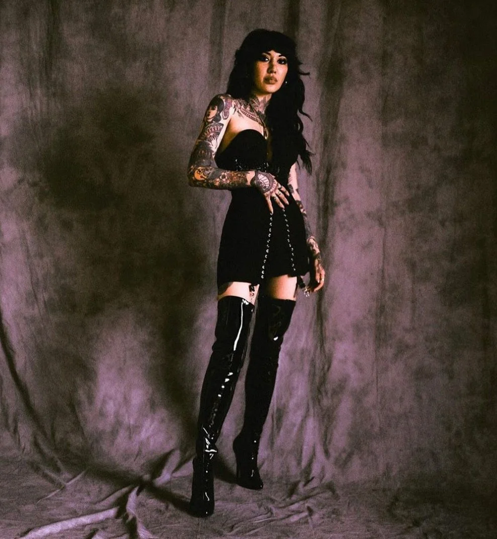 A woman with long black hair, wearing a black dress and thigh-high patent leather boots, stands against a gray textured background. She has tattoos on her arms and holds a chain.