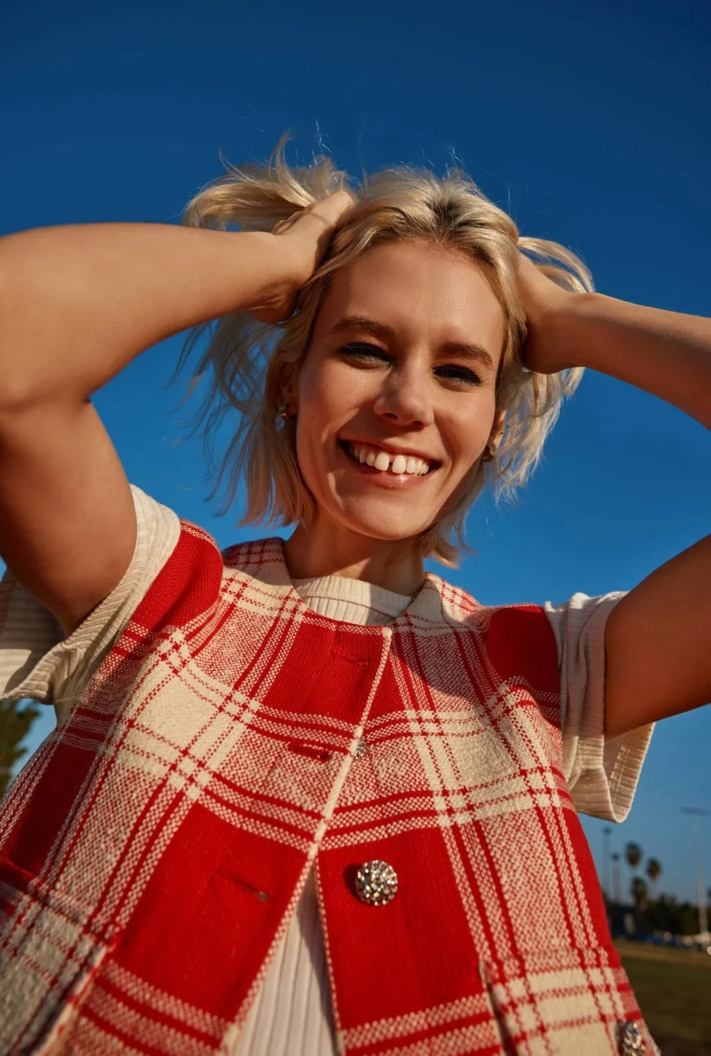 A woman smiling and holding her blonde hair, wearing a red and beige plaid vest with a pearl button, against a clear blue sky.