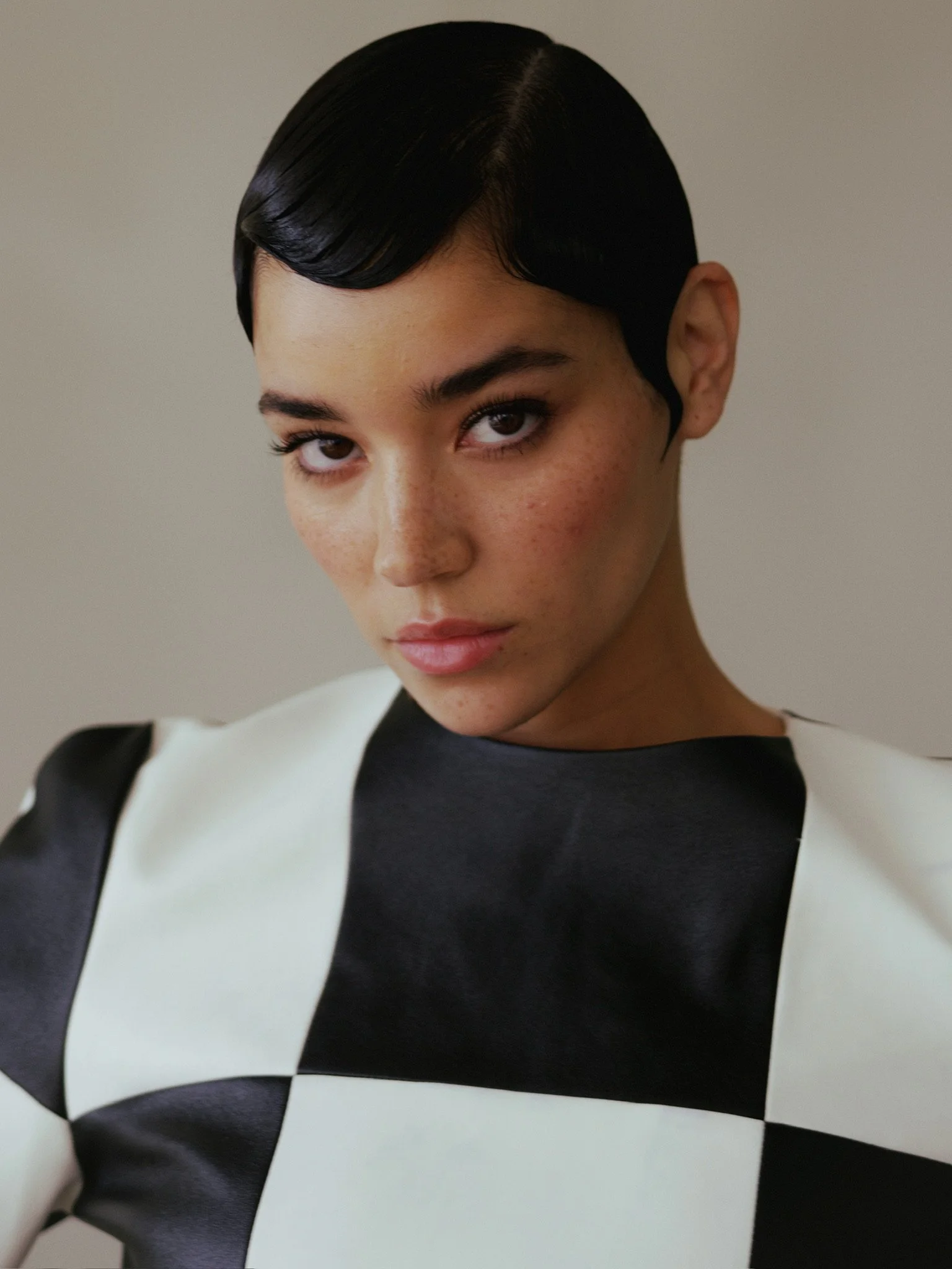 A woman with short black hair styled in waves, wearing a black and white dress, looking at the camera against a plain background.