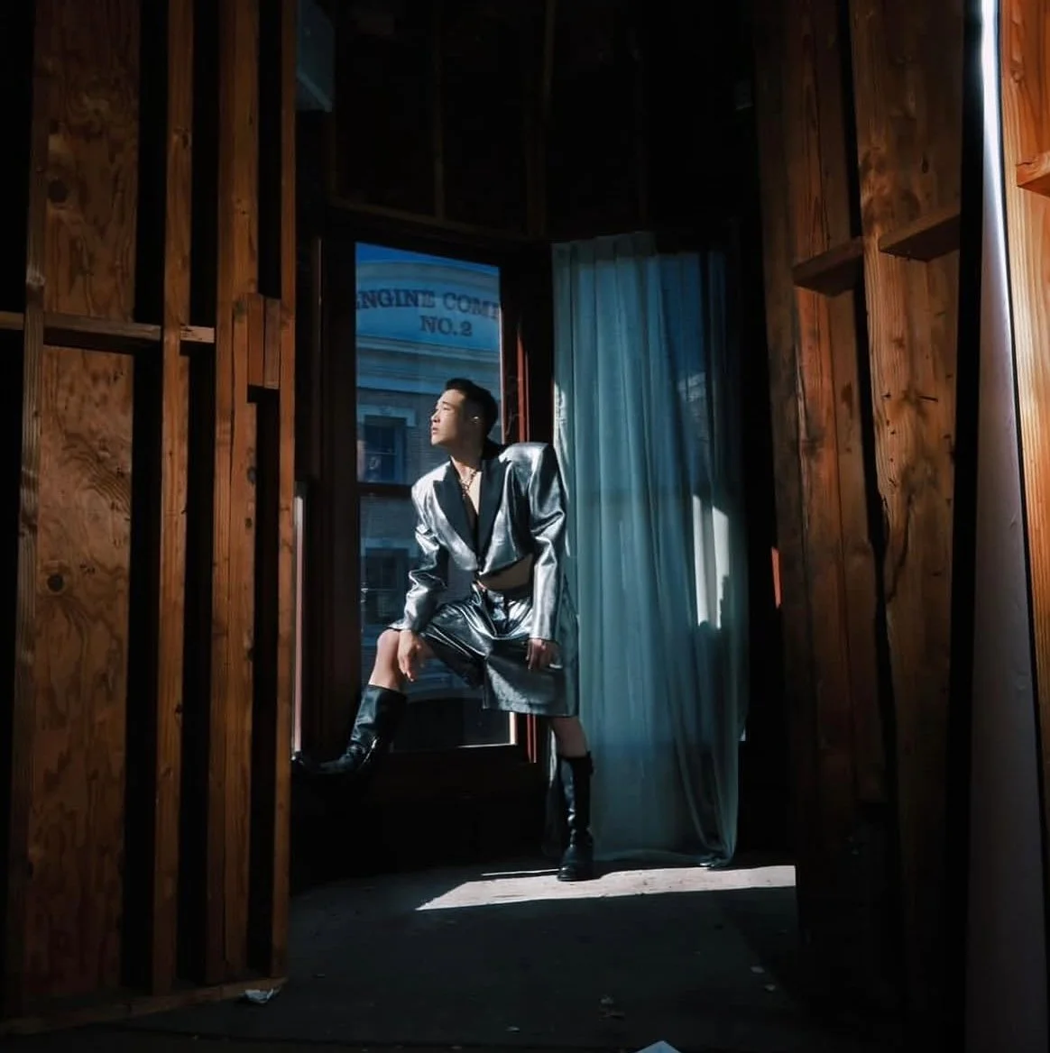 A person dressed in shiny silver and black fashion posing in a wooden room near a glass door with sunlight streaming in.