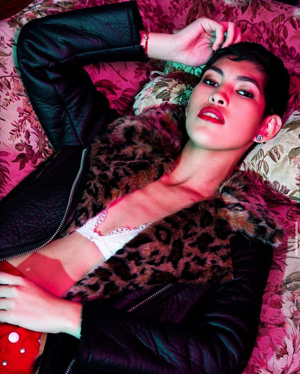 A woman lying on a floral-patterned couch, wearing a black leather jacket with leopard print fur collar, red lipstick, and earrings, with her hand near her head.