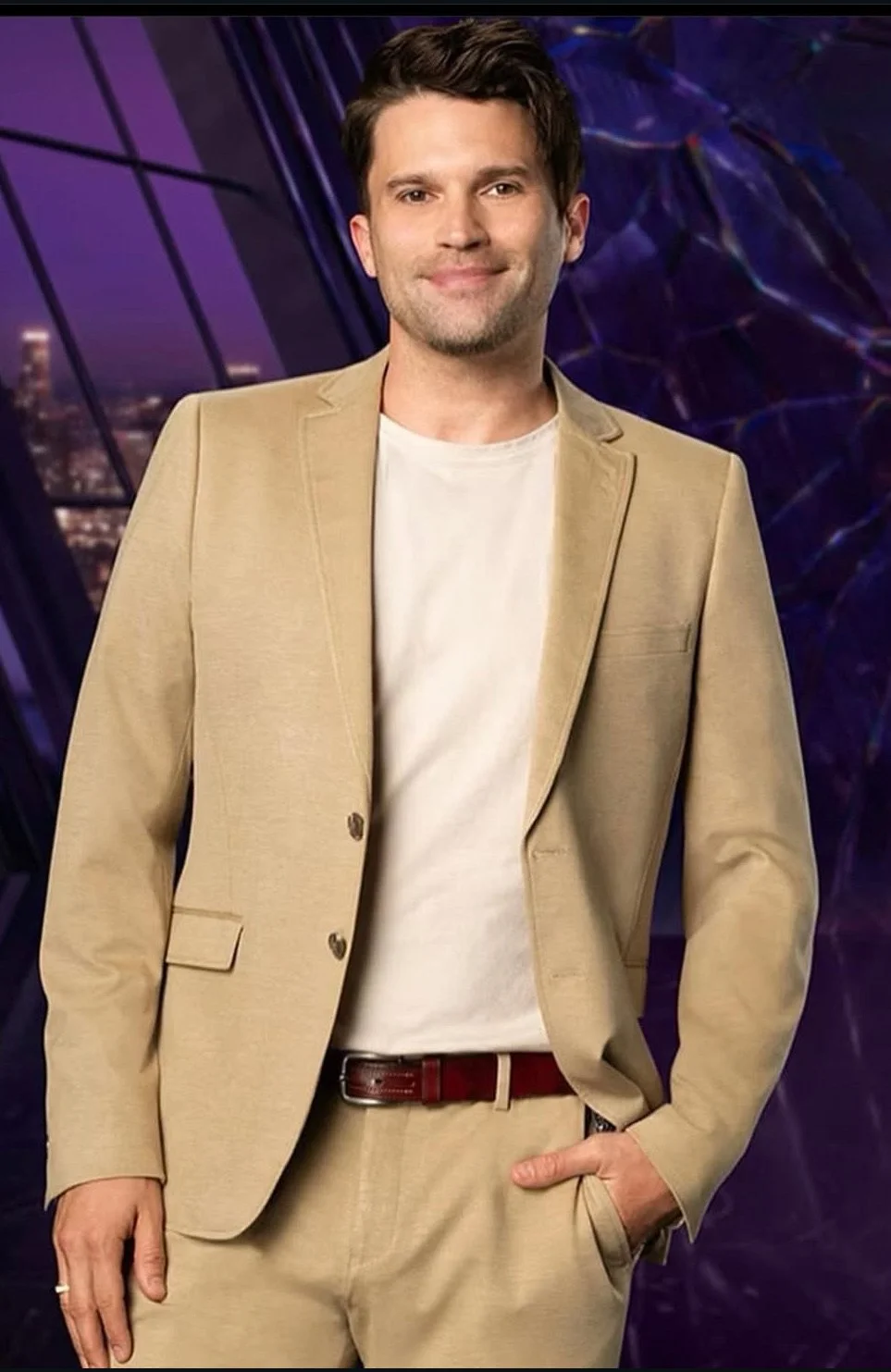 A man with dark hair, dressed in a beige suit and white t-shirt, stands with one hand in his pocket against a purple and black geometric background.