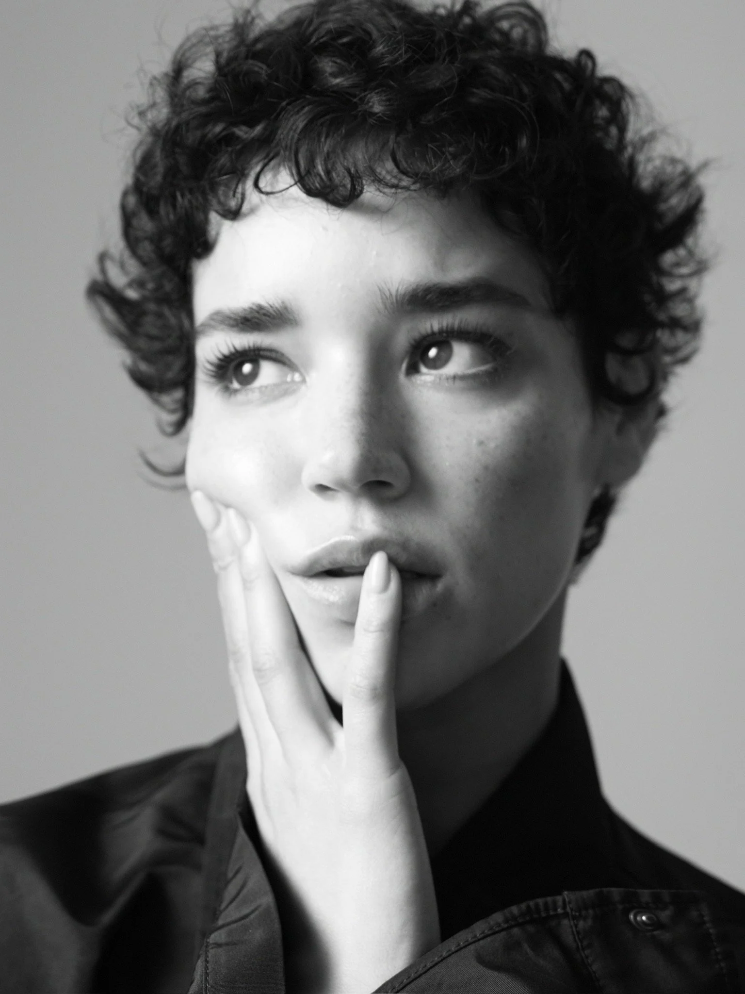 Close-up black-and-white portrait of a woman with curly hair, with her finger resting on her lips, appearing to be thinking or surprised.