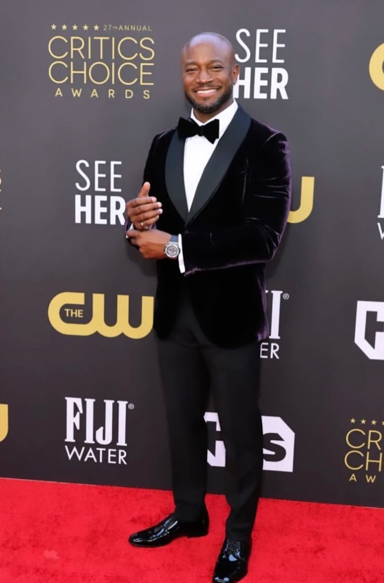 A man in a tuxedo smiling on the red carpet at the Critics Choice Awards. There are logos in the background including Critics Choice Awards, See Her, The CW, FIJI Water, and others.