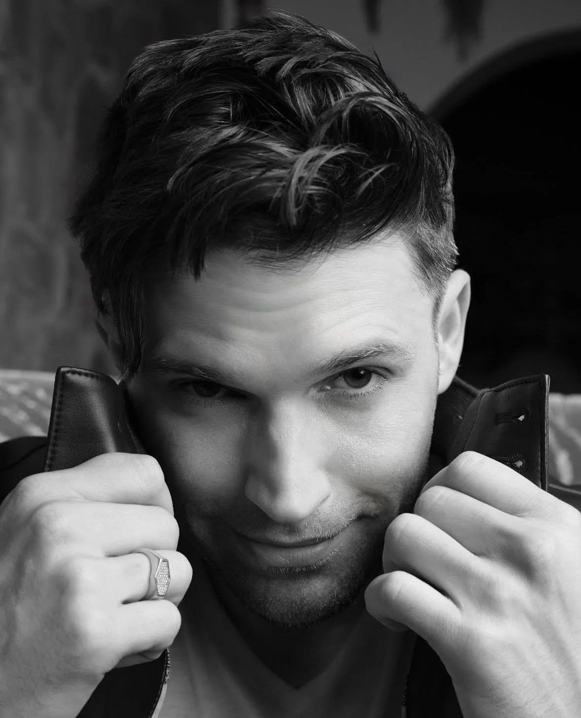 A black and white photo of a young man with short, styled hair, holding the collar of his jacket near his face, looking directly at the camera with a serious expression.