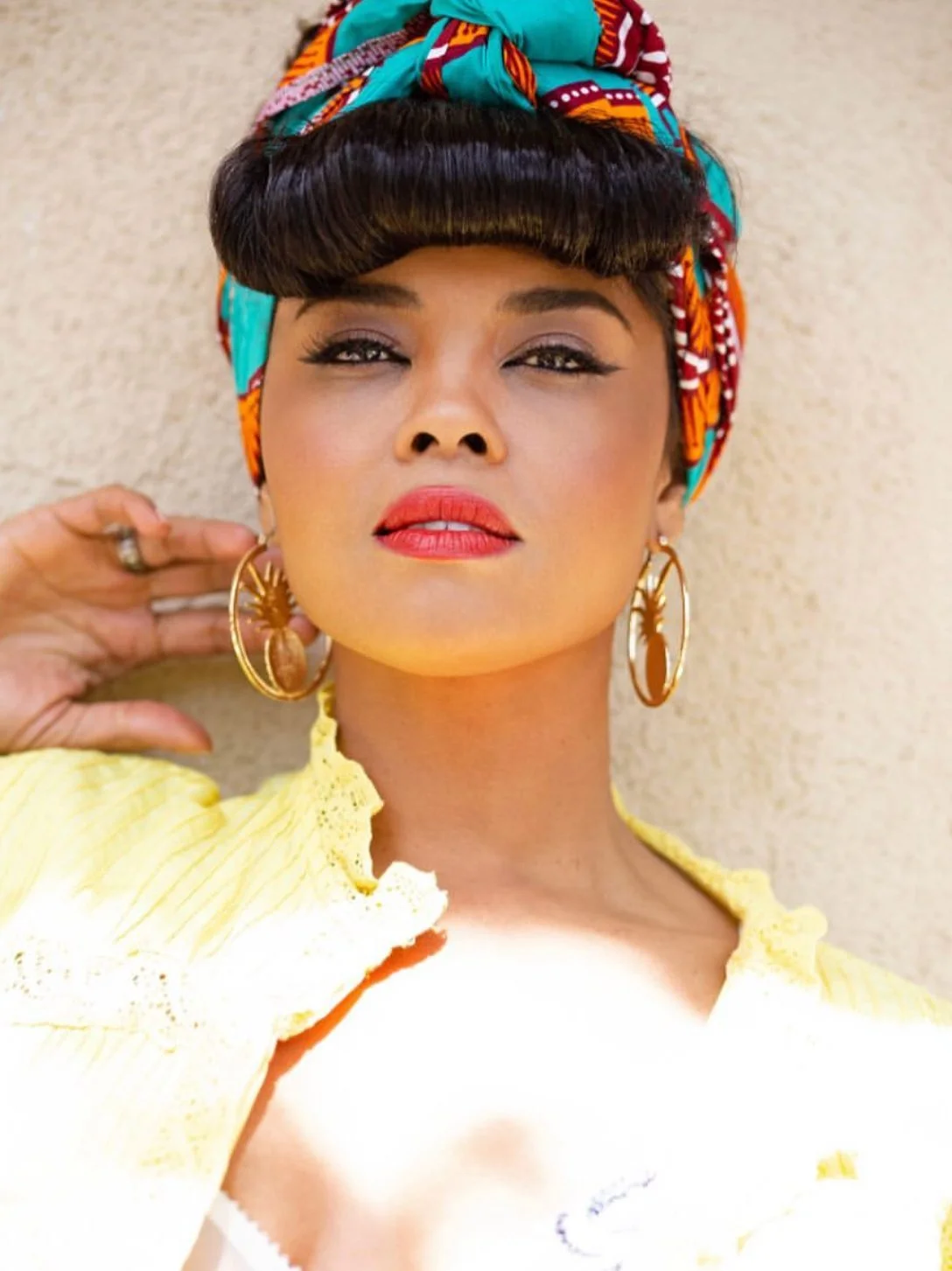 A woman with a colorful headwrap, heavy earrings, and yellow clothing, lying on a beige surface.