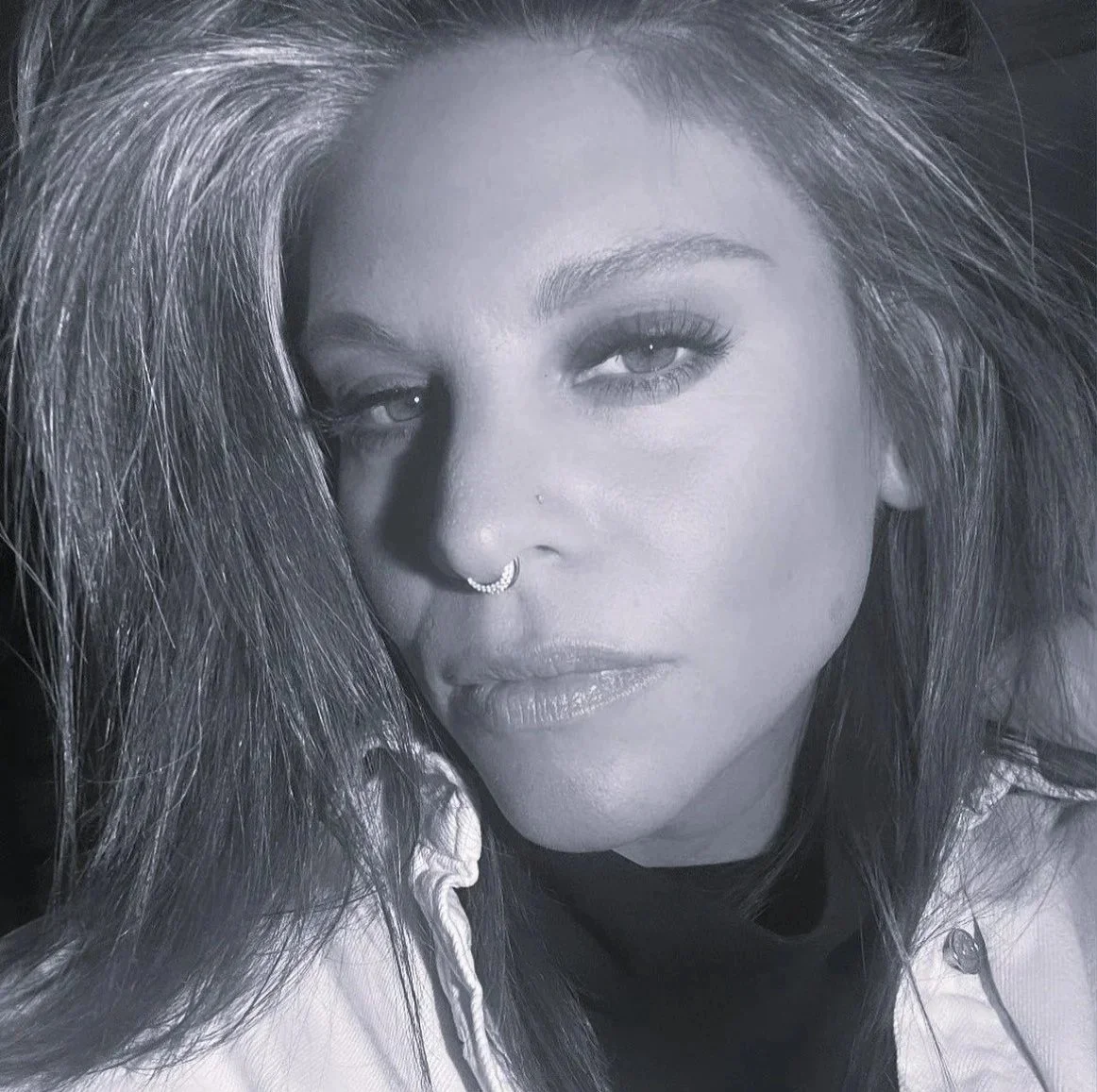 Black and white close-up portrait of a woman with large voluminous hair, wearing a nose ring and a light-colored jacket, looking directly at the camera.