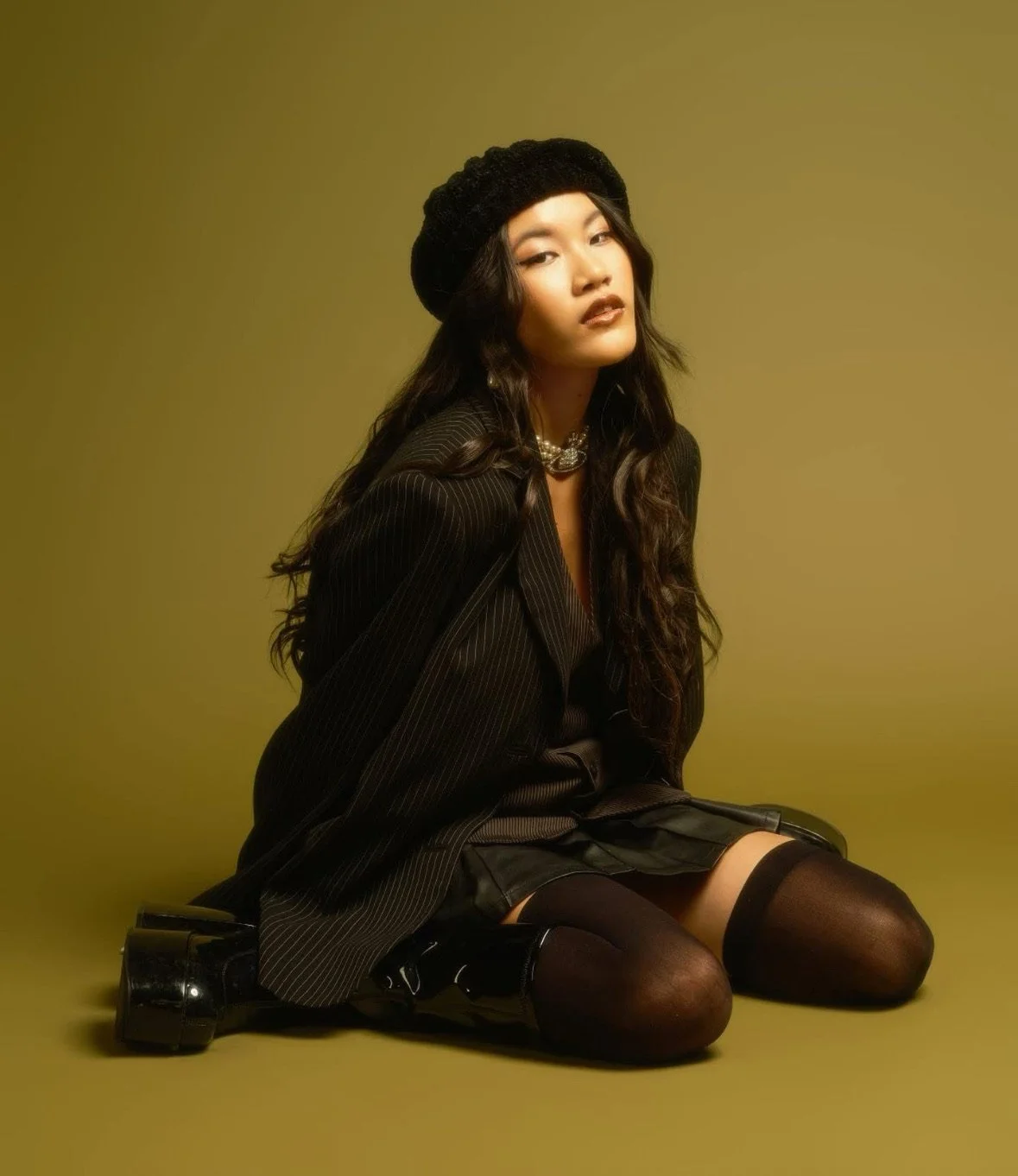 A young woman with long dark hair, wearing a black beret, a pinstripe blazer, mini skirt, thigh-high stockings, and black shiny platform shoes, sitting on the floor against a muted yellow background.