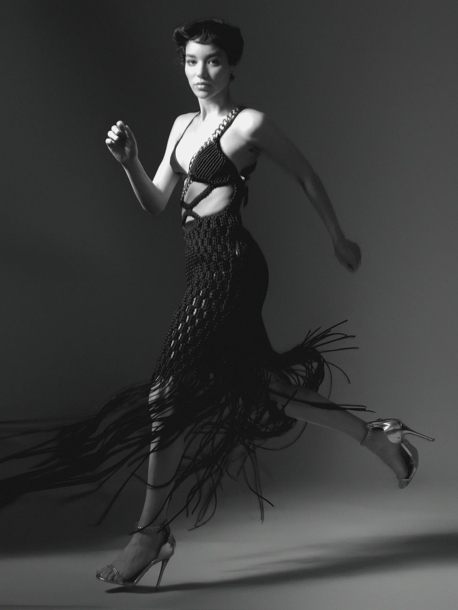 A black-and-white photo of a woman with short hair, wearing a revealing dress with cutouts and high heels, mid-dance pose against a plain background.