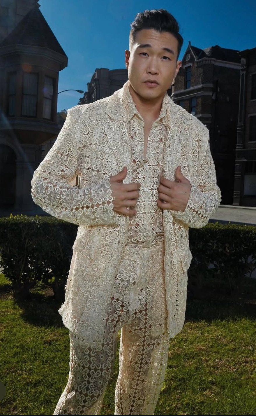 Man standing outdoors in front of Victorian-style houses wearing an intricate cream-colored lace suit.