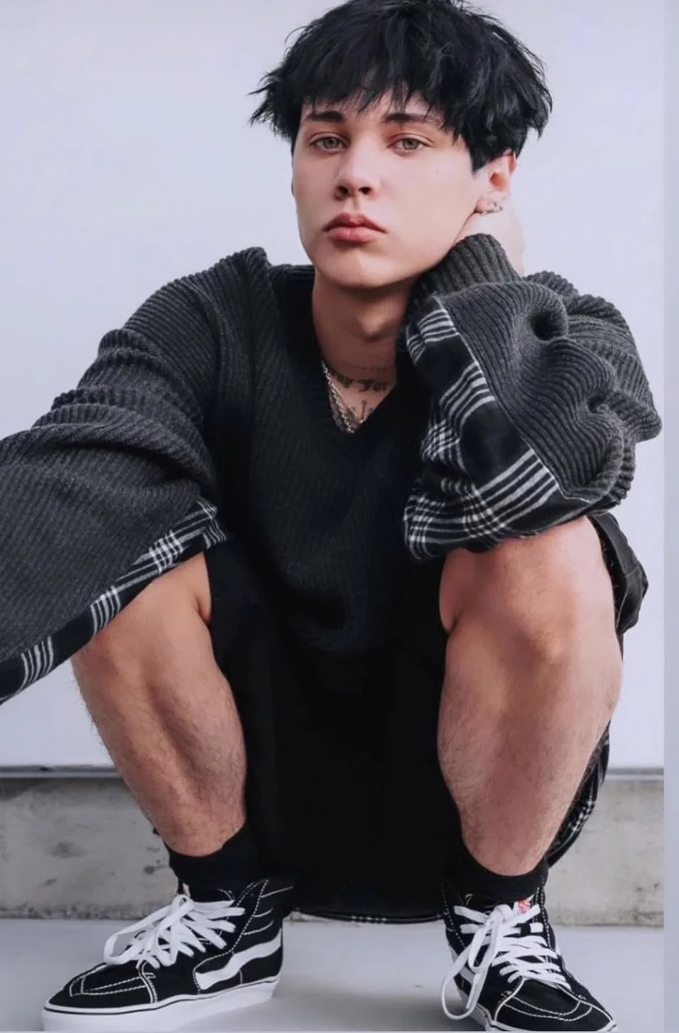 Young person squatting in black sneakers, black shorts, and an oversized black and gray plaid shirt, with a white background.