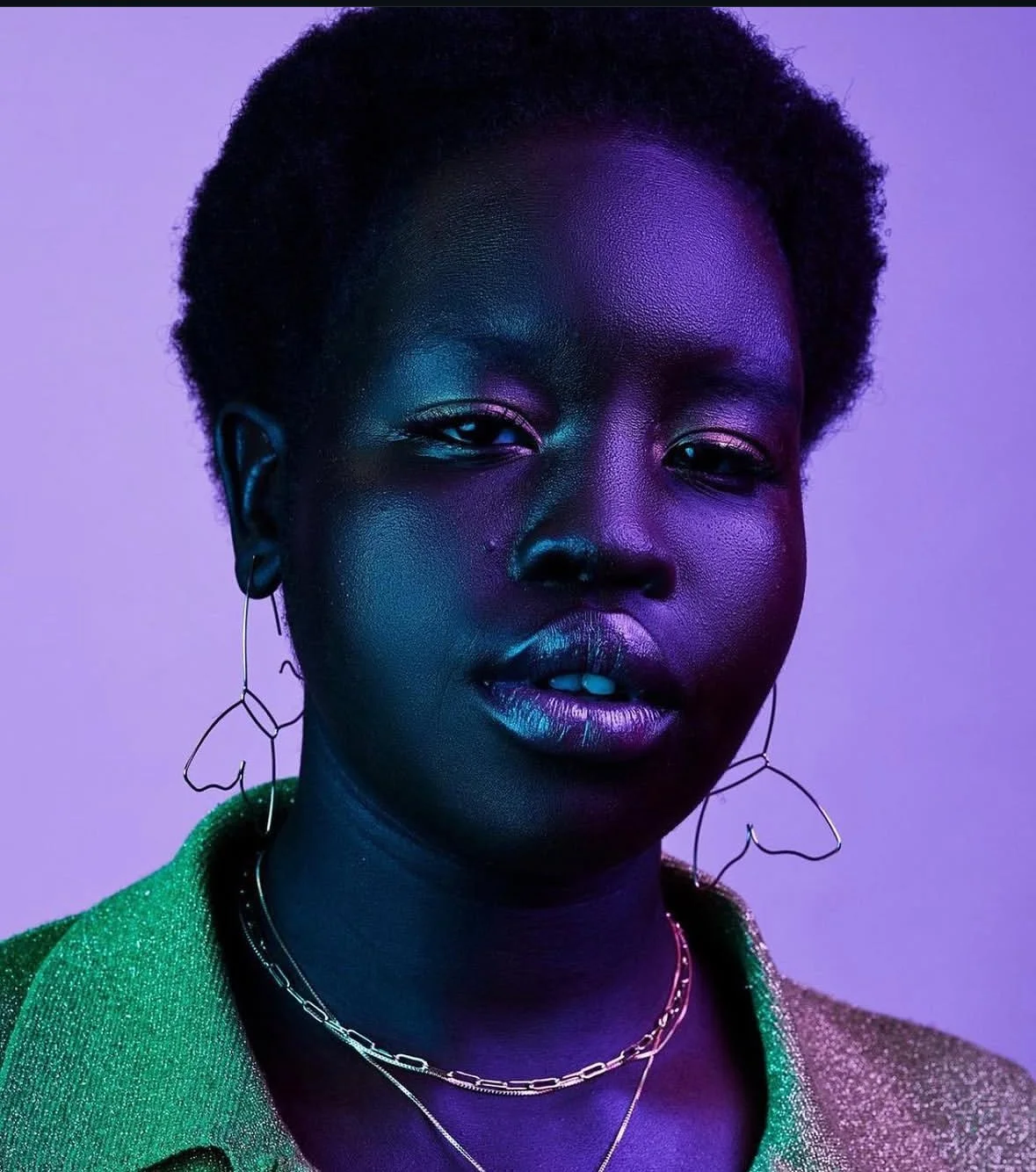 A portrait of a Black woman with short, curly hair, illuminated with purple, blue, and green lighting. She is wearing large butterfly-shaped earrings and layered necklaces, with a green, glittery jacket.