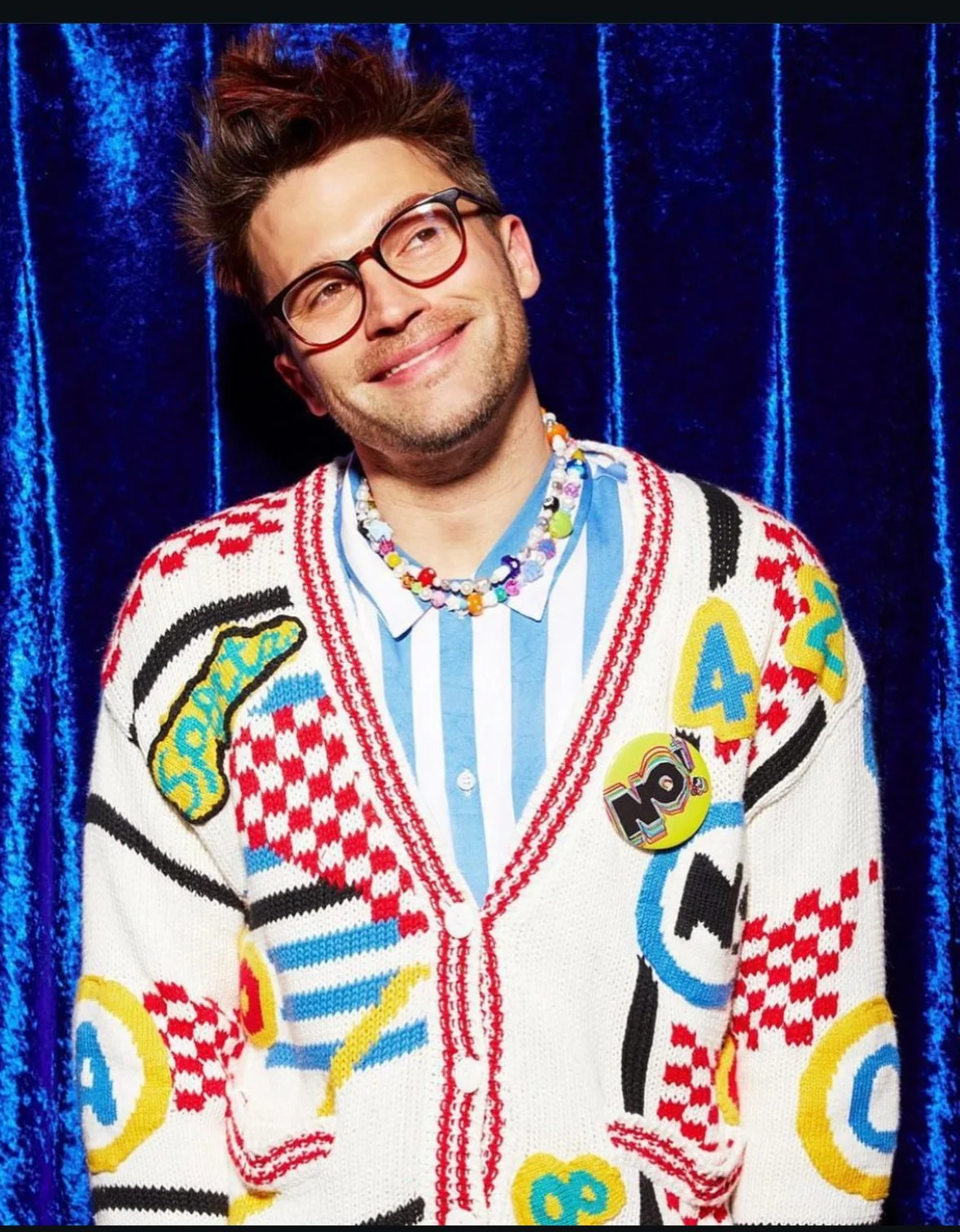 A man smiling in front of a deep blue curtain, wearing glasses, a colorful cardigan with numbers and letters, a pin that says 'WOW!', a layered beaded necklace, and a blue and white striped shirt.