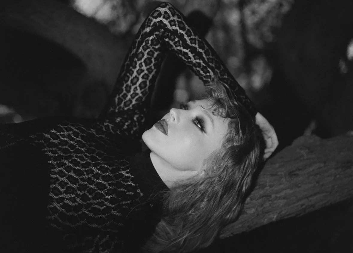 A woman with curly hair lying on a tree trunk, looking upwards in black and white. She wears a patterned long-sleeve top.