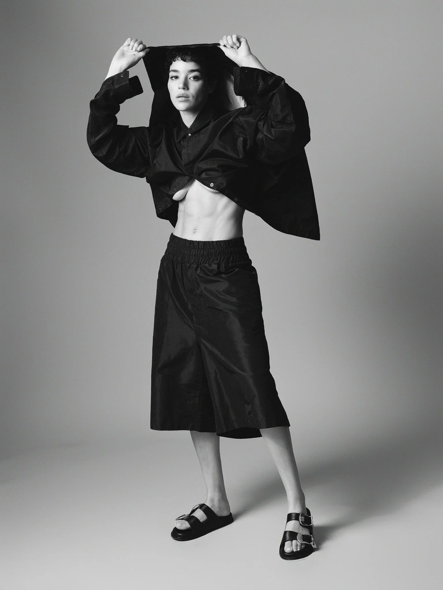 Black and white photo of a woman with curly hair wearing a black shirt and wide pants, lifting her shirt to reveal her midriff. She is wearing sandals with buckles and standing against a plain background.