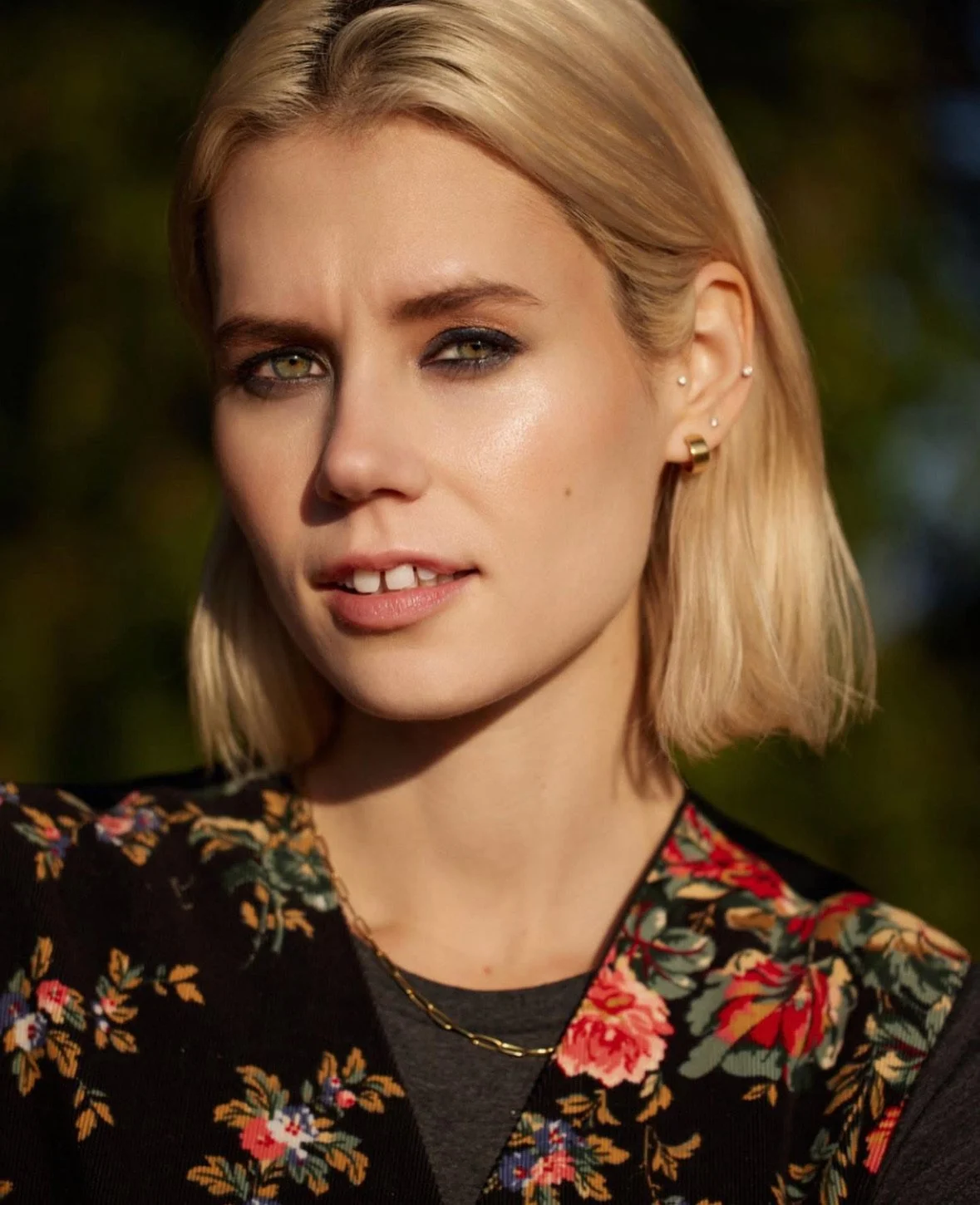 Portrait of a young woman with blonde hair and light makeup, wearing a floral patterned top and gold jewelry, outdoors during daytime.