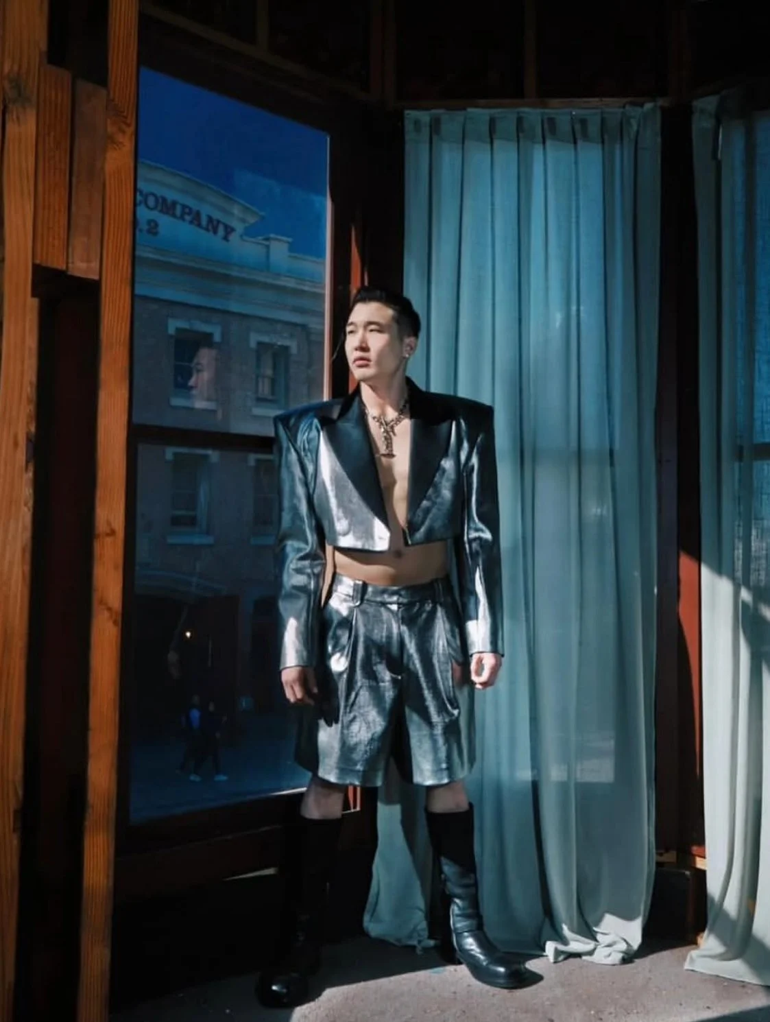 Person wearing a shiny silver and black jacket, matching shorts, and black knee-high boots standing next to a window with curtains.