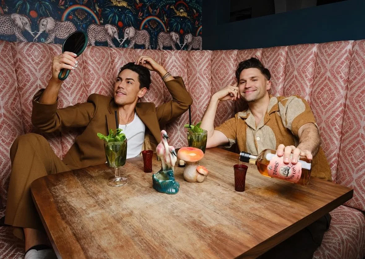 Two men sitting at a wooden table in a restaurant with pink patterned booth seating. One man on the left, dressed in a brown suit, is holding a mirror and smiling. The other man on the right, dressed in a short-sleeve, color-blocked shirt, is pouring