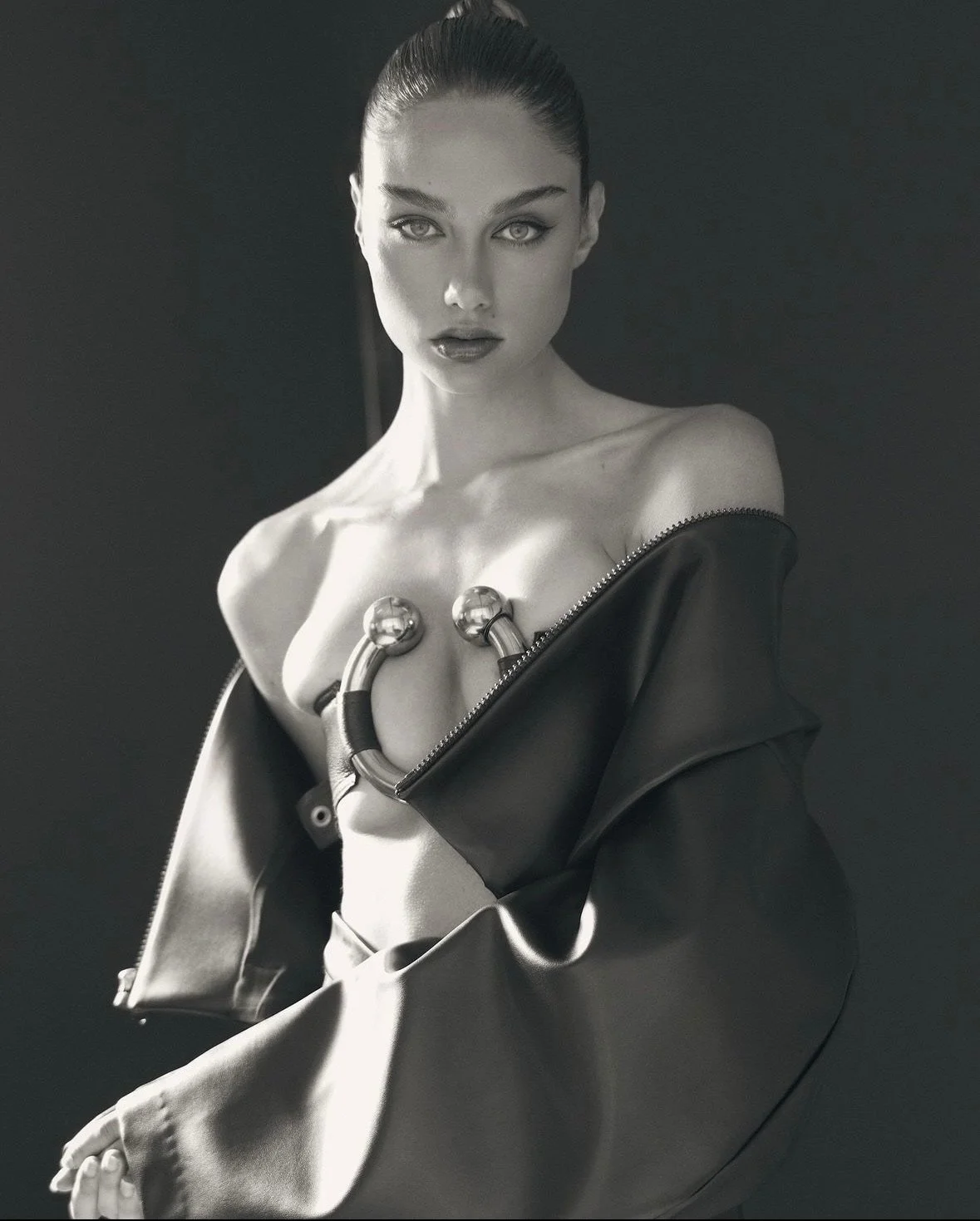 Black and white photo of a woman with slicked-back hair, looking directly at the camera, partially covered by a leather jacket hanging off her shoulders, with metallic tubes around her chest as a fashion statement.