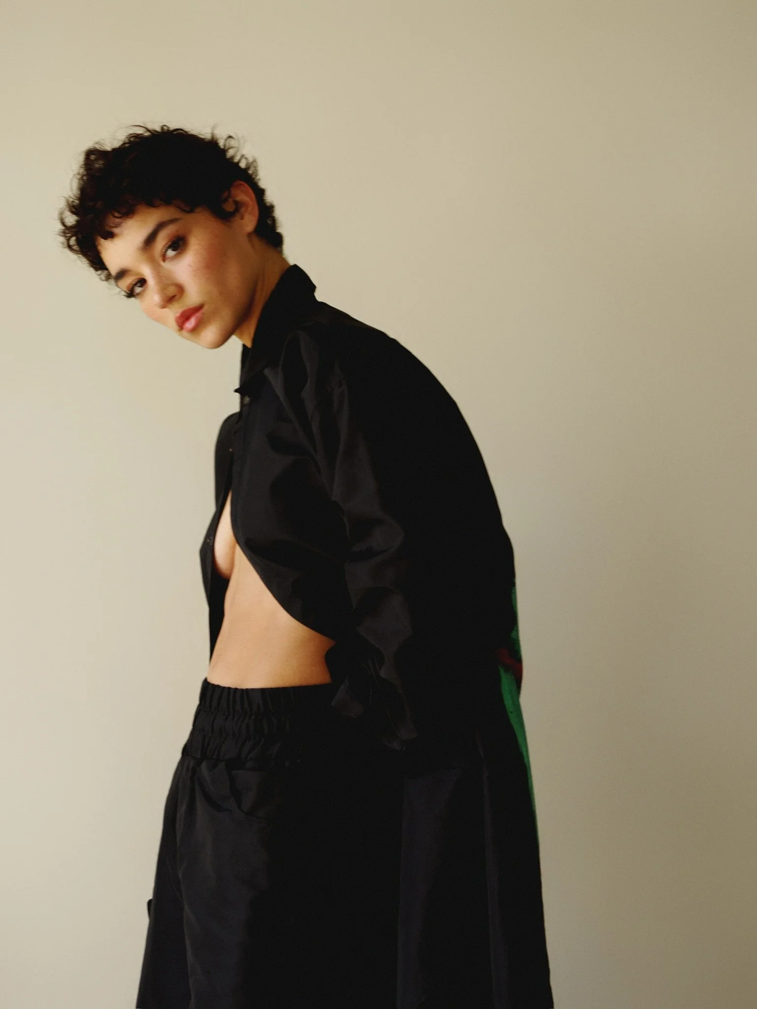 A young woman with short curly dark hair, wearing a black jacket and shorts, standing against a plain beige wall.