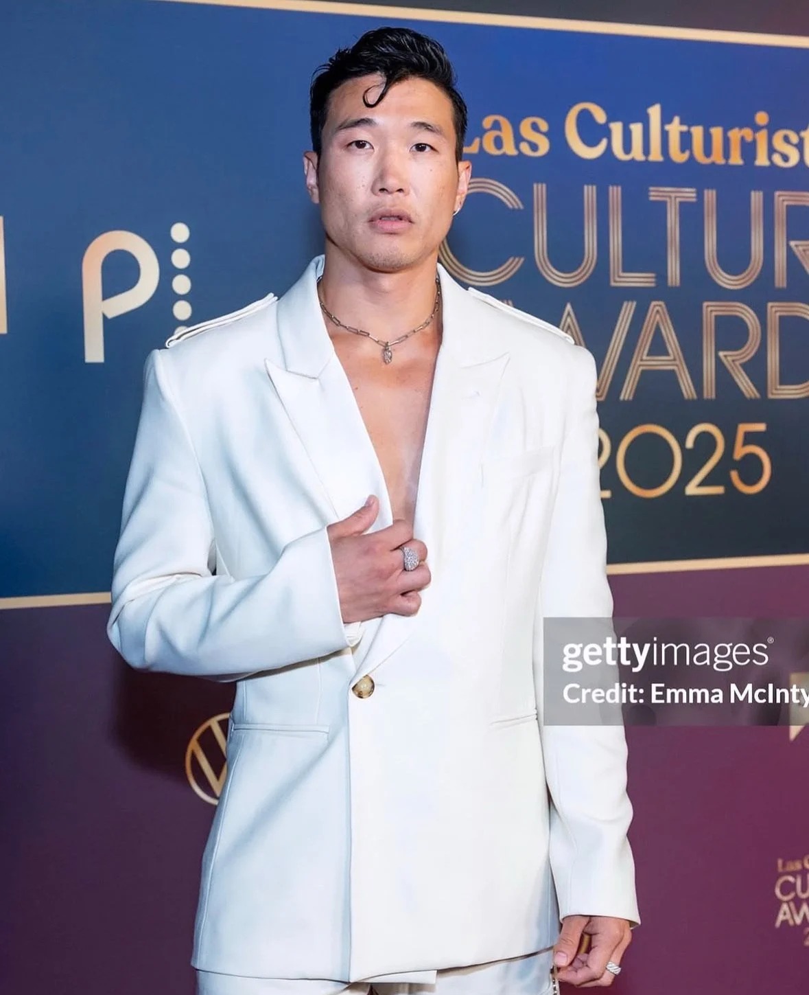 Man in a white suit with jewelry posing on red carpet at Las Cultures Award 2025 with blue background.