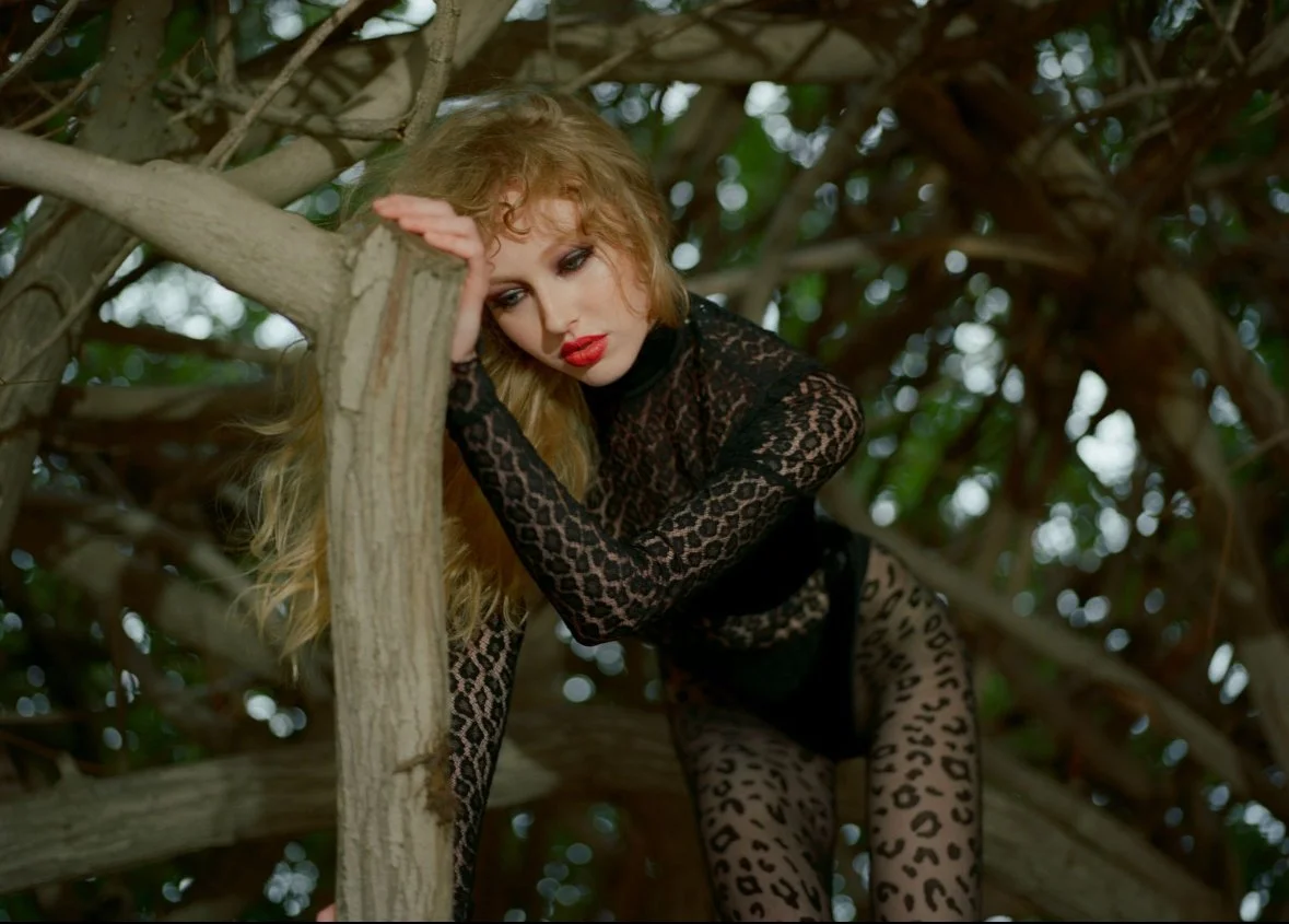 A woman dressed in black lace and animal print lingerie, leaning on a tree branch, surrounded by dense foliage.
