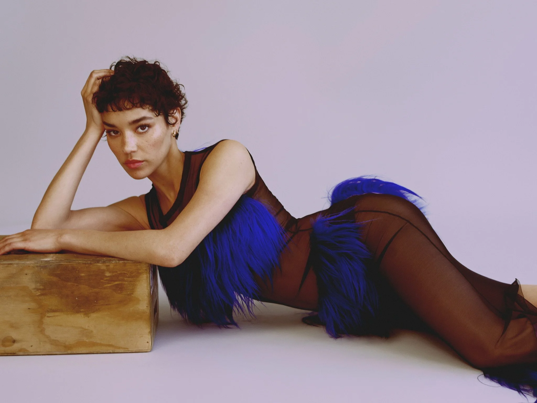 A woman with short curly brown hair, looking directly at the camera, lying on her side with her elbow resting on a wooden surface, wearing a sheer black outfit with blue feathers at the waist, against a plain light-colored background.