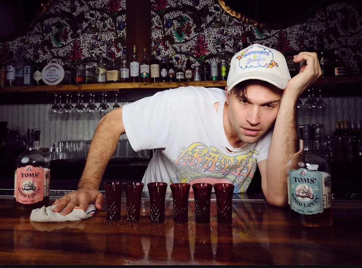 A young man with a distressed expression leaning on a bar counter, wearing a white T-shirt with holes and a white cap with colorful embroidery. In front of him are six red shot glasses, two bottles of Toms tequila, and a small towel. The background f