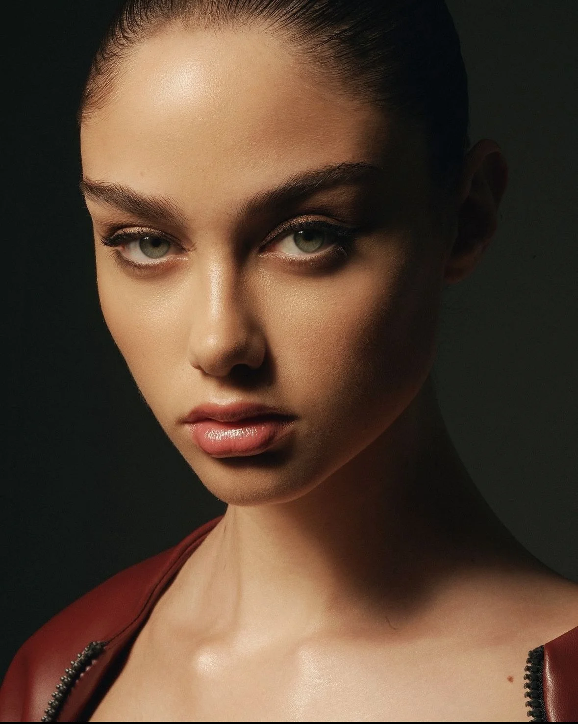 Close-up of a woman with blonde hair tied back, green eyes, and natural makeup, wearing a red leather jacket with a zipper, posed against a dark background.