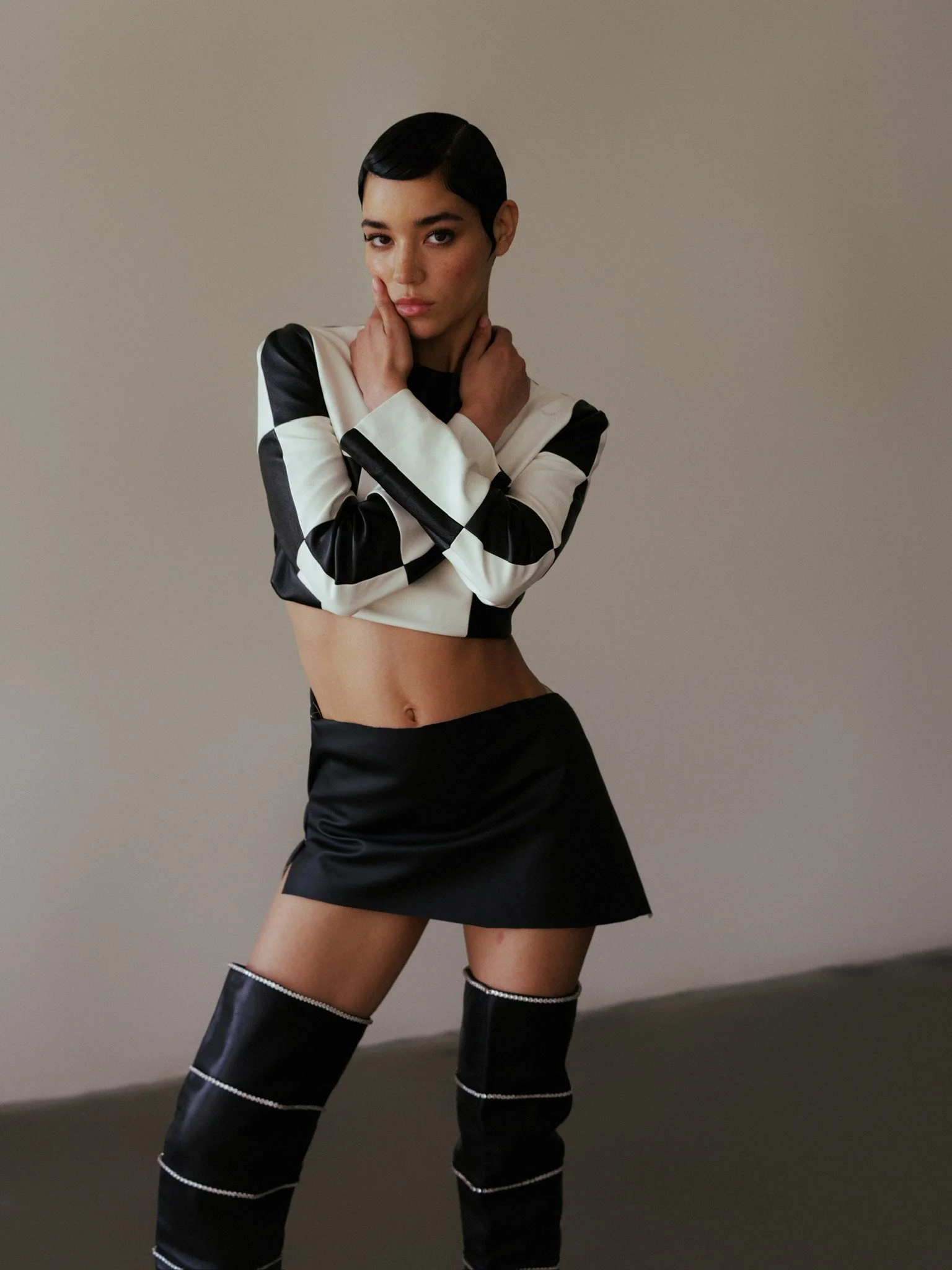 A woman with short dark hair and a serious expression, wearing a black and white checkered cropped jacket, black shorts, and thigh-high black boots with silver accents, stands against a plain gray background.