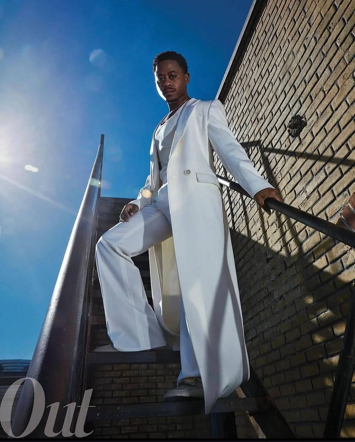 A man in a white suit standing on outdoor metal stairs next to a brick wall, under a bright blue sky.