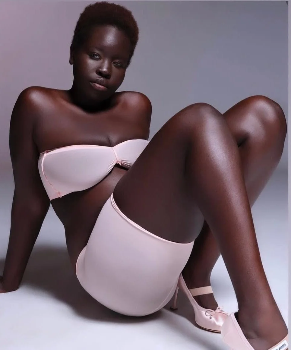 A woman with dark skin and short hair, wearing pale pink lingerie and matching high heels, sitting on the floor with her knees bent and looking at the camera.
