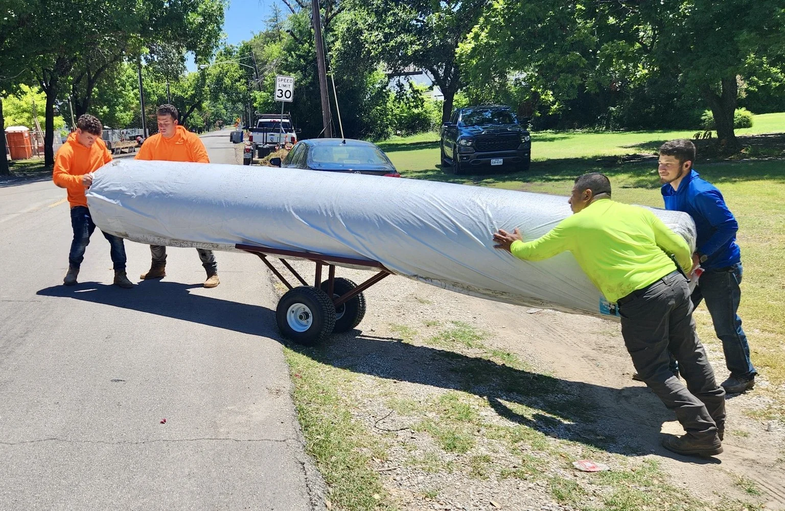 Four workers maneuver a large carpet roll supported by the two-wheeled Carpet Dolly on a residential street.