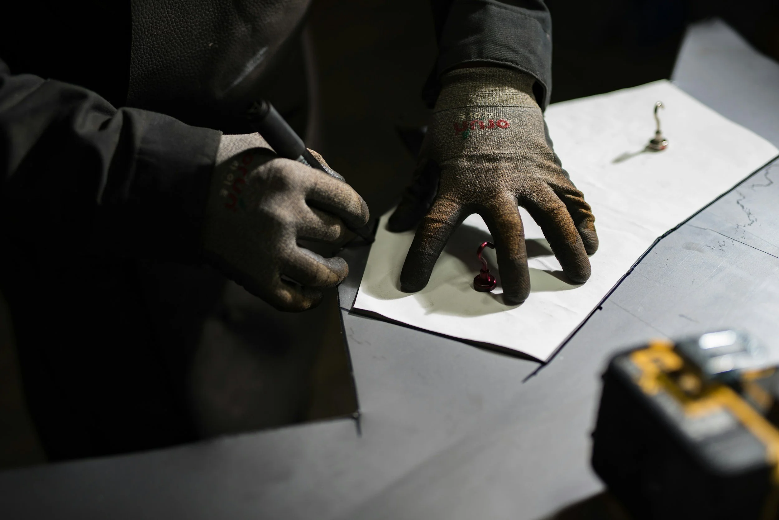 Manos con guantes trabaja en un proyecto, usando un rotulador para marcar un patrón en papel, con agujas de coser y una caja de herramientas visible.