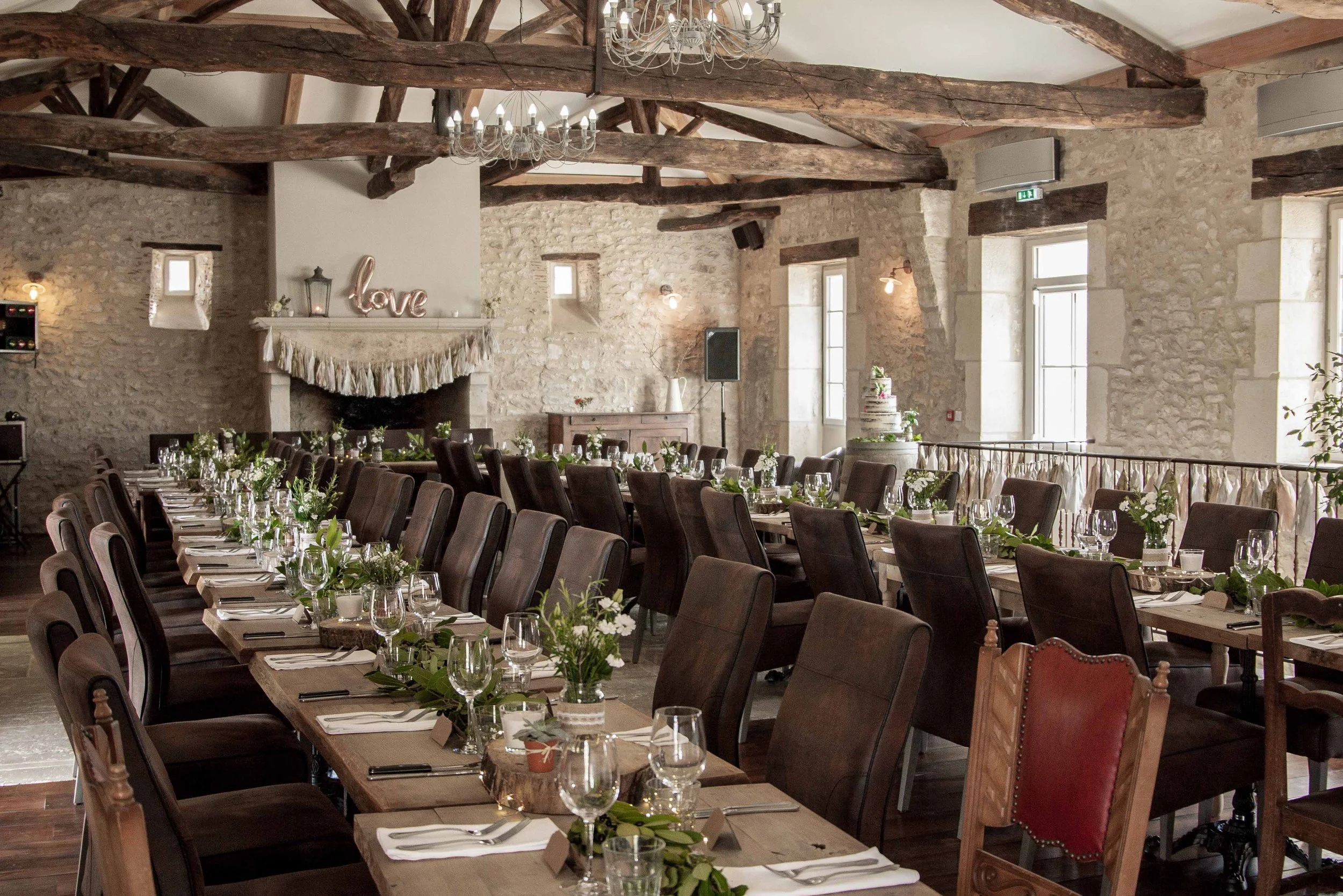 A rustic wedding reception setup inside a stone building with wooden beams on the ceiling. Long tables are decorated with greenery and glassware, and there are chairs with various styles around the tables. Chandeliers hang from the ceiling, and a fireplace with a sign that says 'love' is visible in the background.