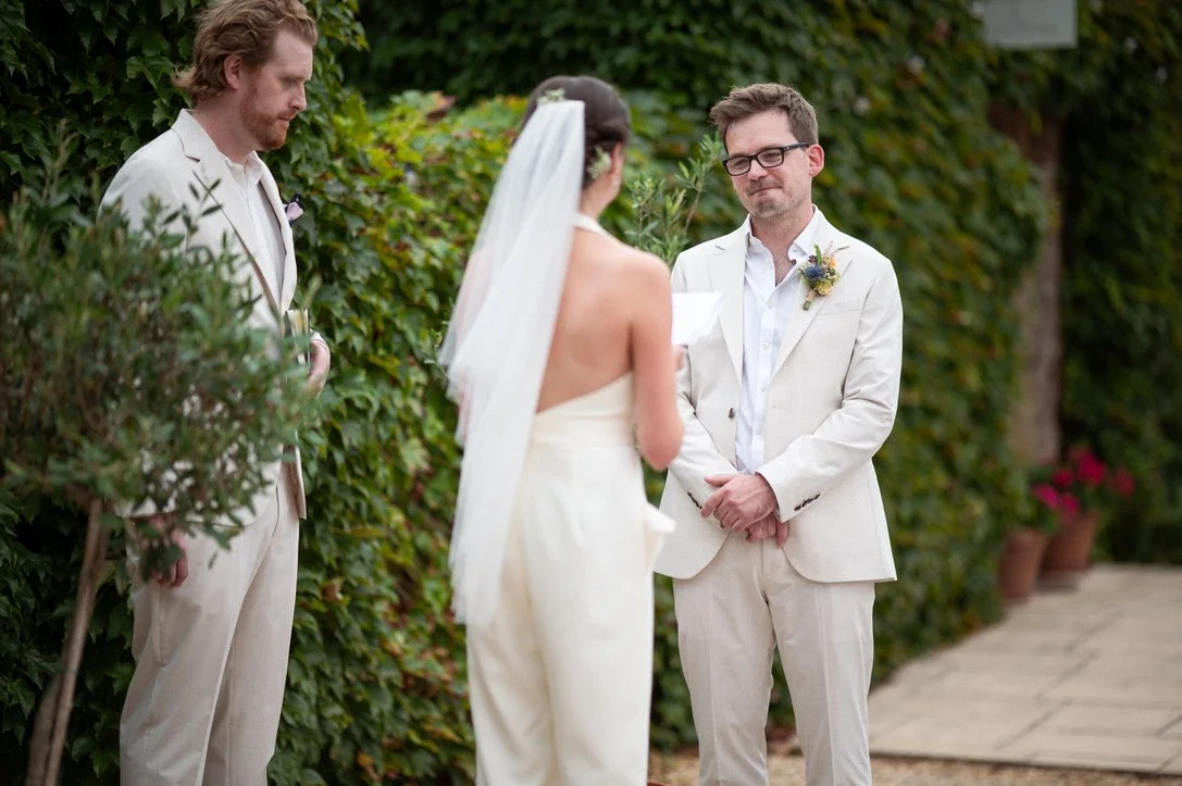 A woman in a strapless wedding dress with a veil is reading a marriage vow in front of two men in white suits, outdoors with green foliage in the background.