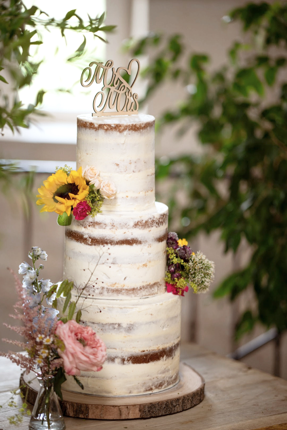 A three-tier semi-naked wedding cake decorated with fresh flowers, including sunflowers, pink roses, and other colorful blooms, topped with a wooden 'Mr & Mrs' cake topper, placed on a wooden cake board surrounded by additional flower arrangements.