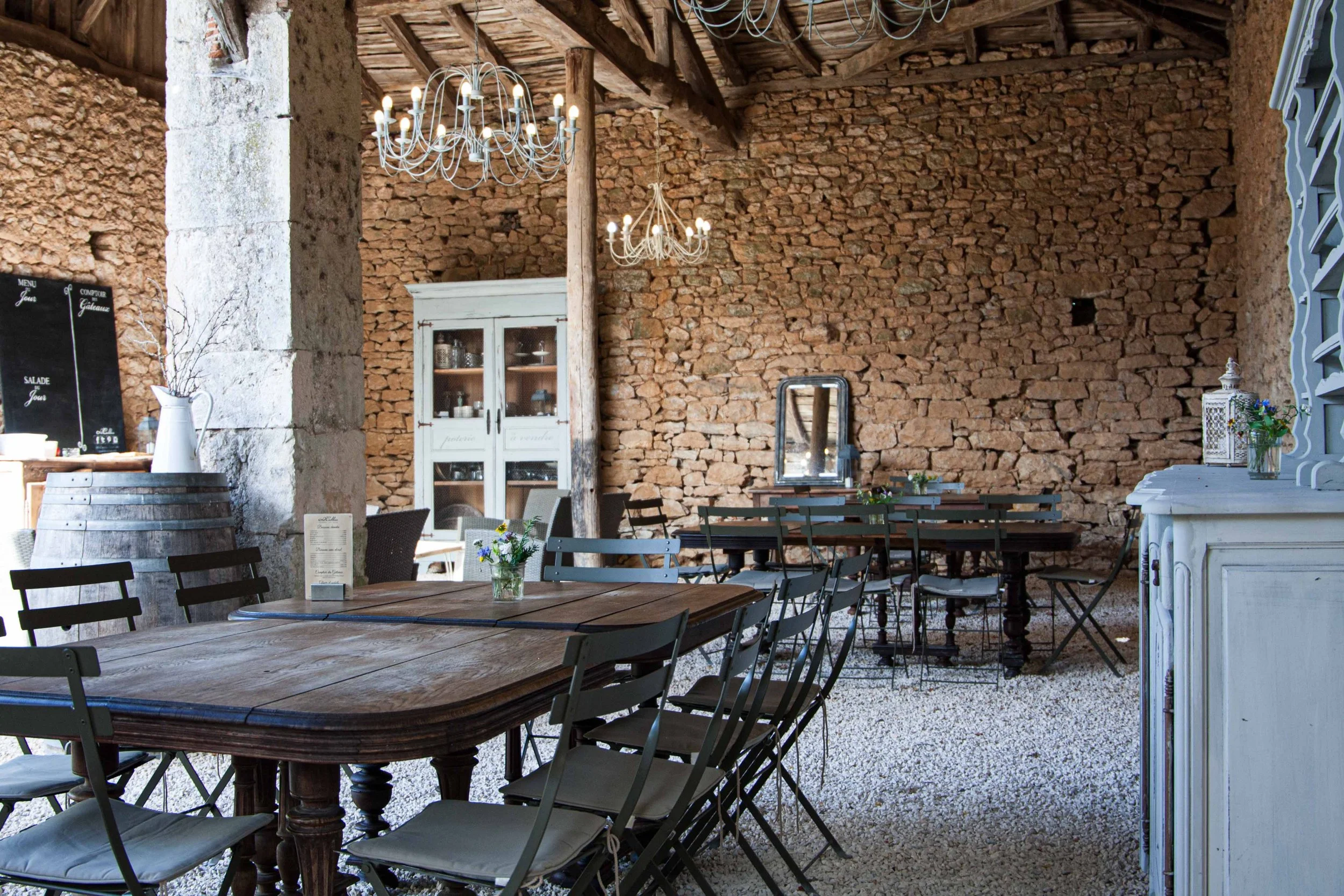 Interior of a rustic restaurant or café with exposed brick walls, wooden beams, chandeliers, and tables with chairs and small flower arrangements.