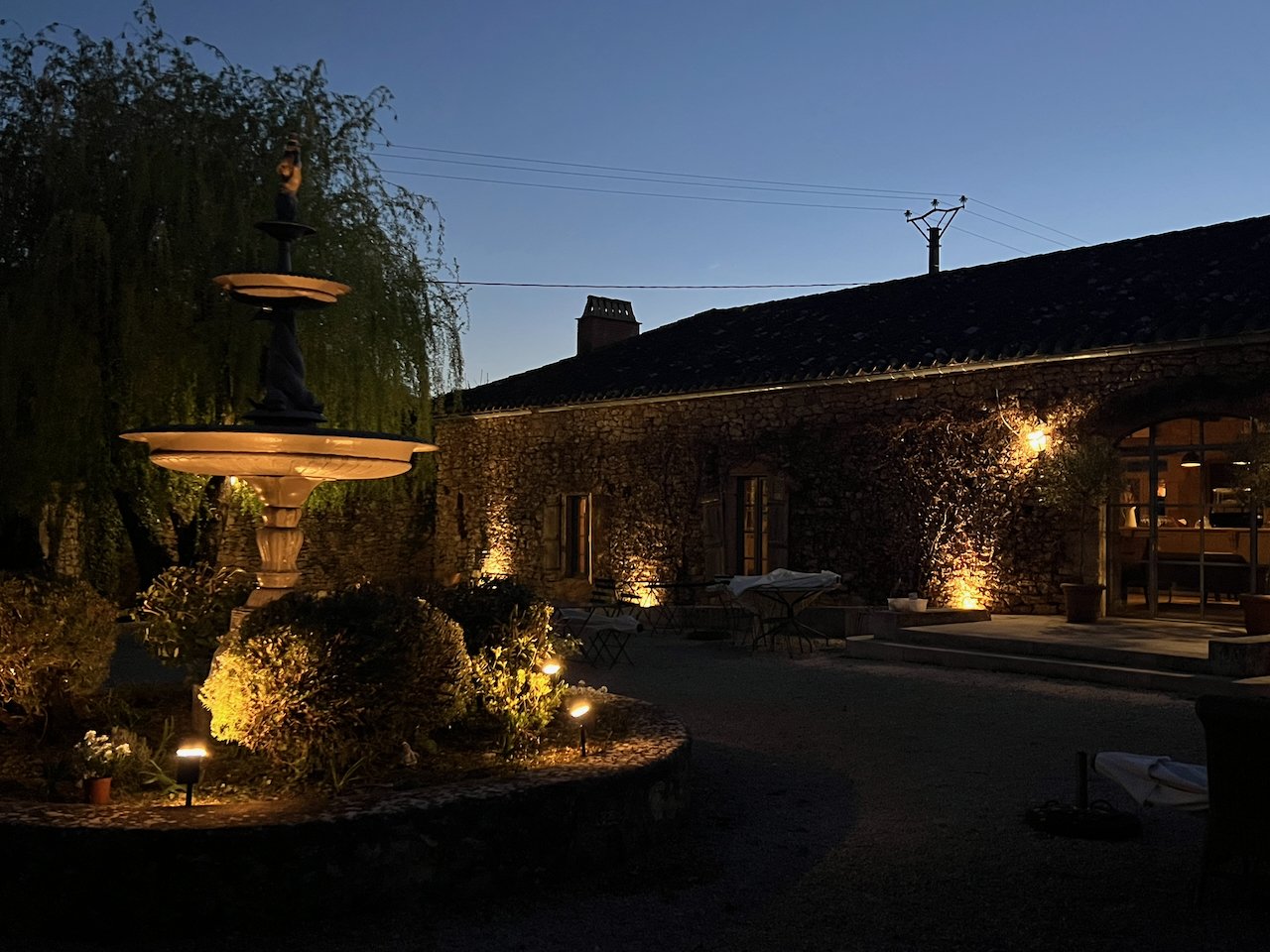 Evening outdoor scene of a rustic stone building illuminated by exterior lights, with a fountain, garden, and patio furniture under a clear sky.