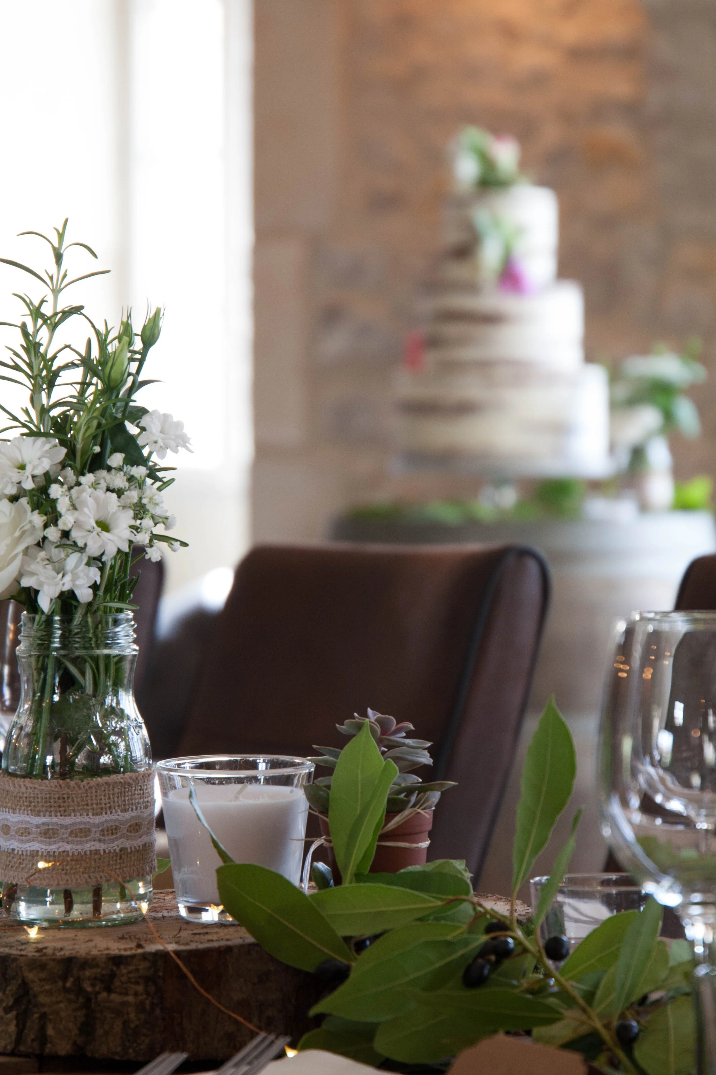 A table decorated with a bouquet of white flowers in a jar wrapped with lace, surrounded by green leaves and small black berries, with a candle in a glass holder and other plants nearby. In the background, a multi-tiered white cake with floral decorations is visible on a stand, blurred against a brick wall and a light source.