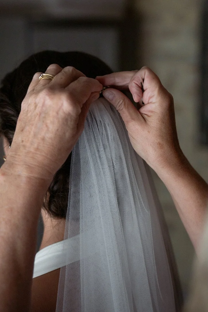 Close-up of hands fastening a bridal veil to a bride's hair, showing the back of the bride's head and the texture of the veil.
