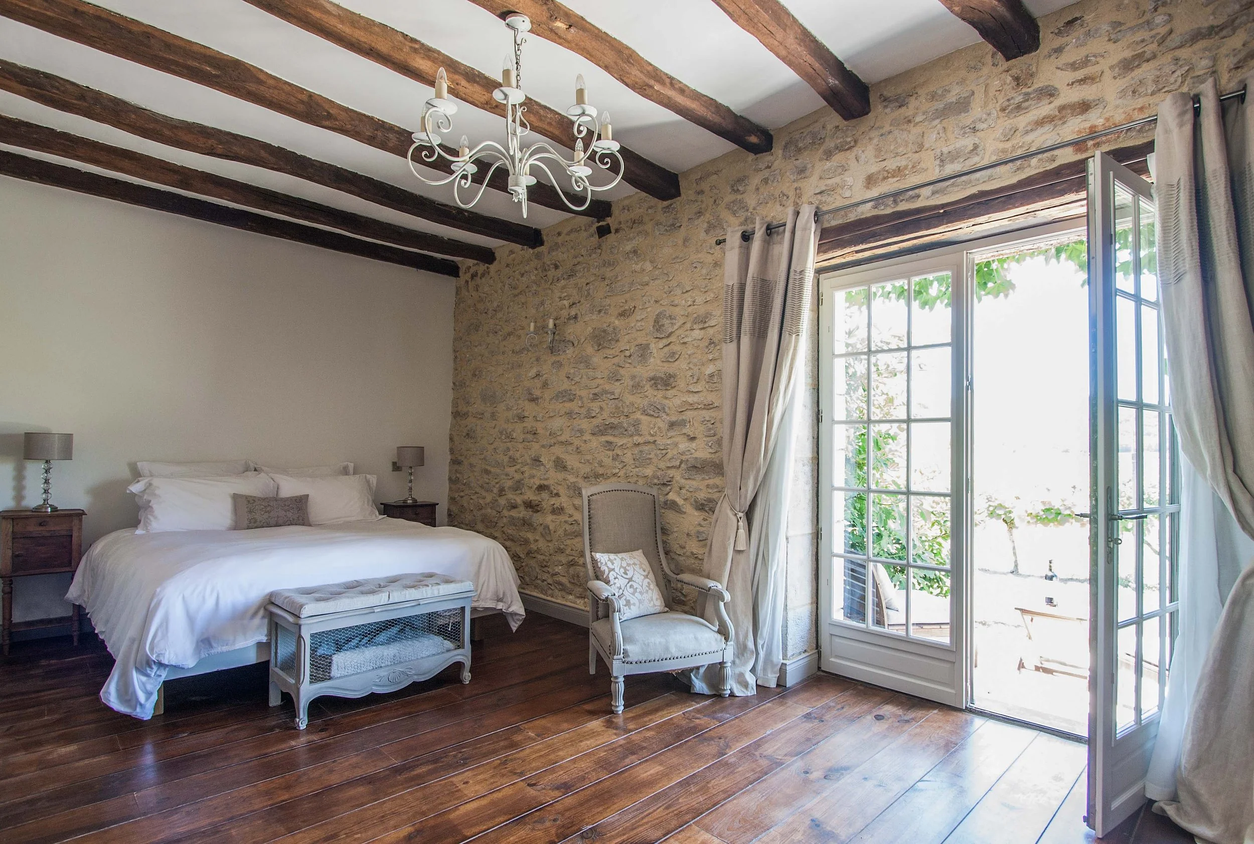 A bedroom with a rustic interior featuring exposed wooden beams on the ceiling, a stone accent wall, a large window with curtains, a white chandelier, a bed with white bedding, two side tables with lamps, and a beige upholstered chair.