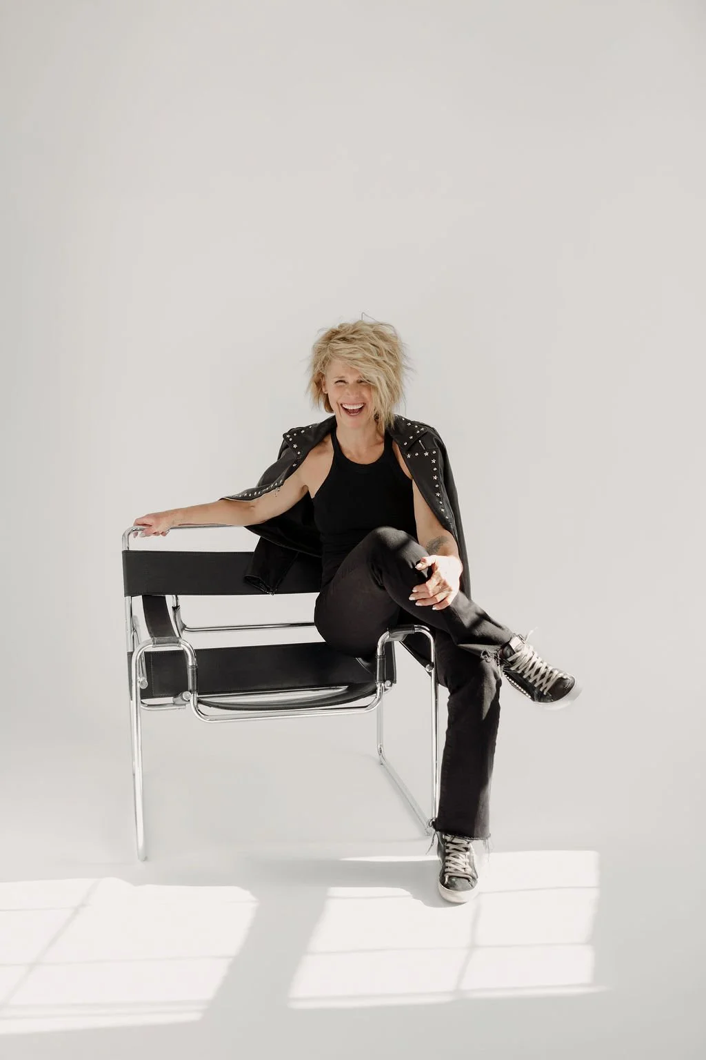 Woman with blond, tousled hair sitting on a modern black and chrome chair, laughing and wearing a black sleeveless top, black pants, and a black leather jacket with metal embellishments.